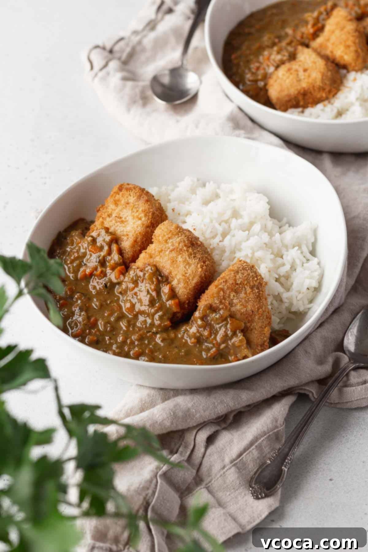 Vegan katsu curry in a white bowl on a linen with a flat leaf parsley plant in the corner of the shot.