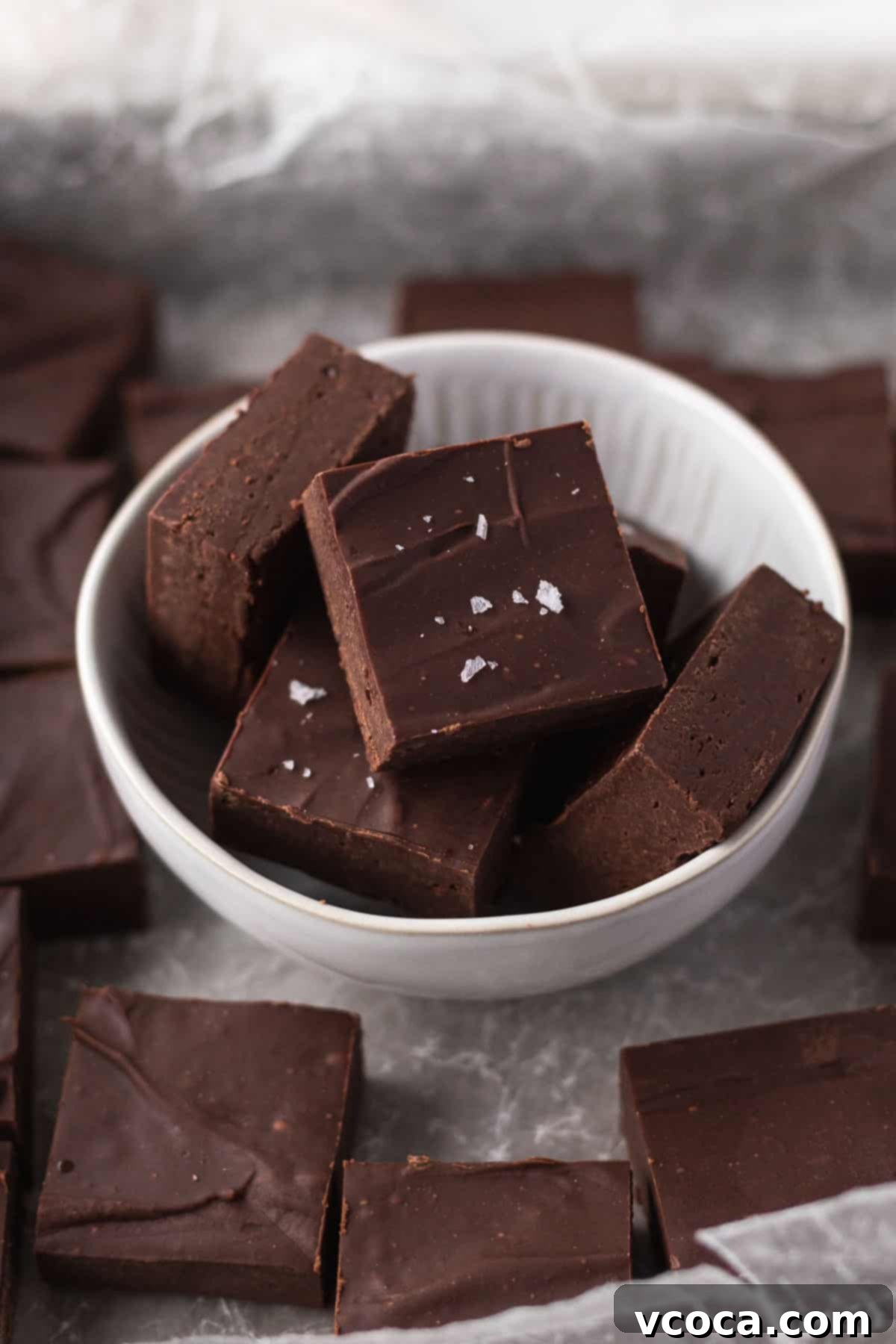 Plant-Based Fudge 2 45 degree angle shot of cubes of dairy-free fudge in a white bowl on a piece of crumpled white parchment with other cubes of fudge.