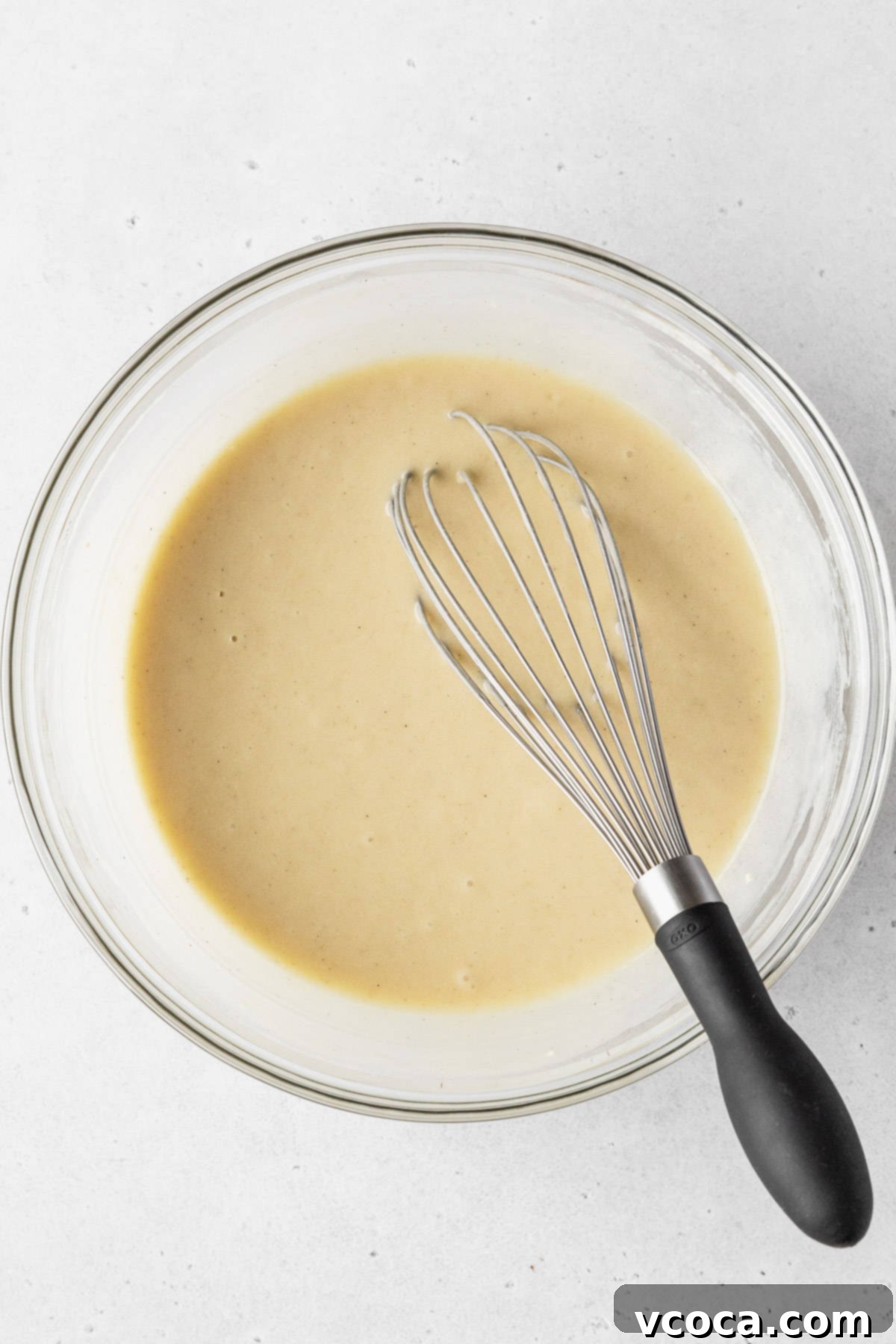Completed dairy free vanilla cupcake batter in a clear mixing bowl with a black-handled whisk.