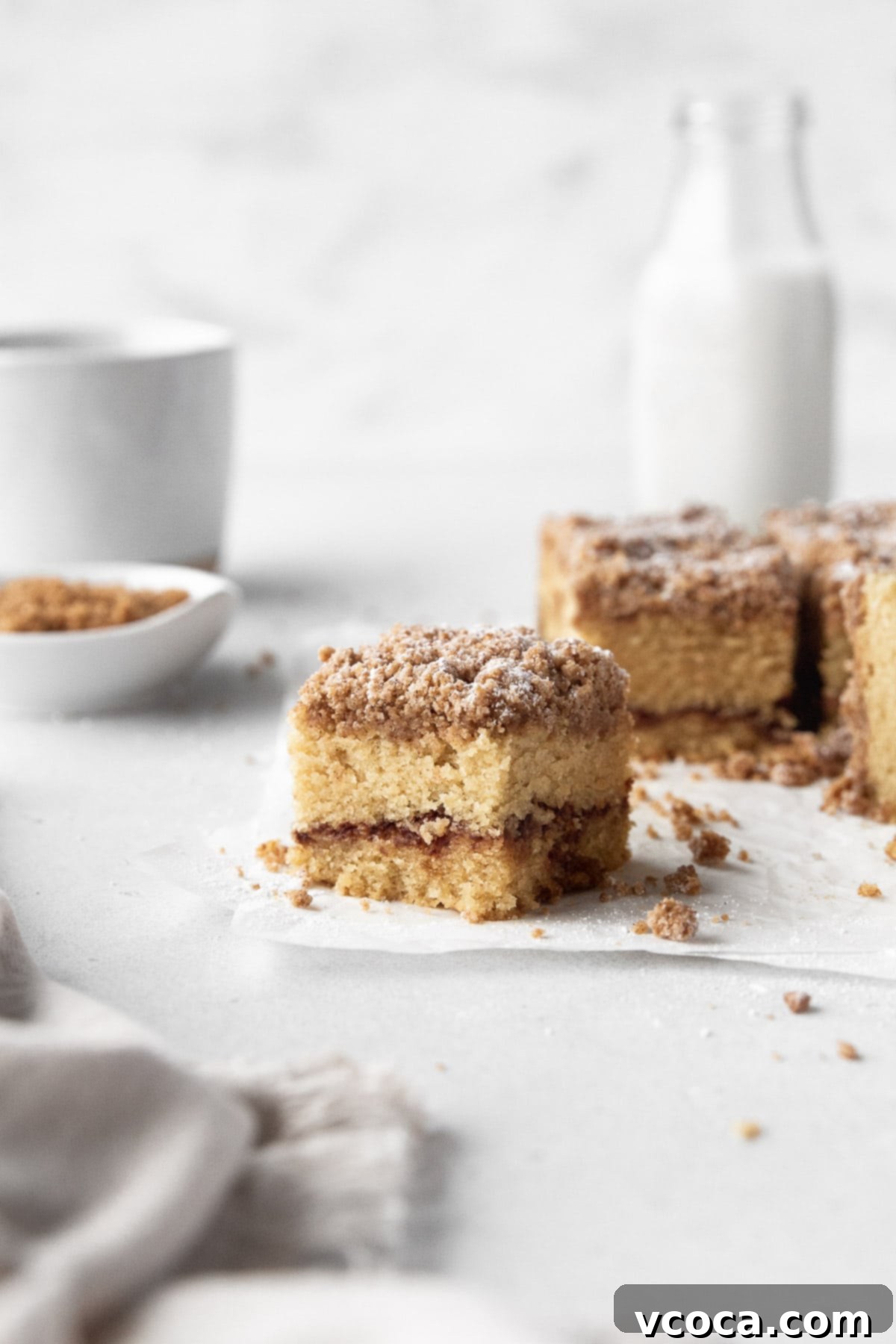 Deliciously Guilt-Free Coffee Cake 13 Square of GF cinnamon crumble cake with the remaining cake, extra crumble topping, and a jug of milk in the background.