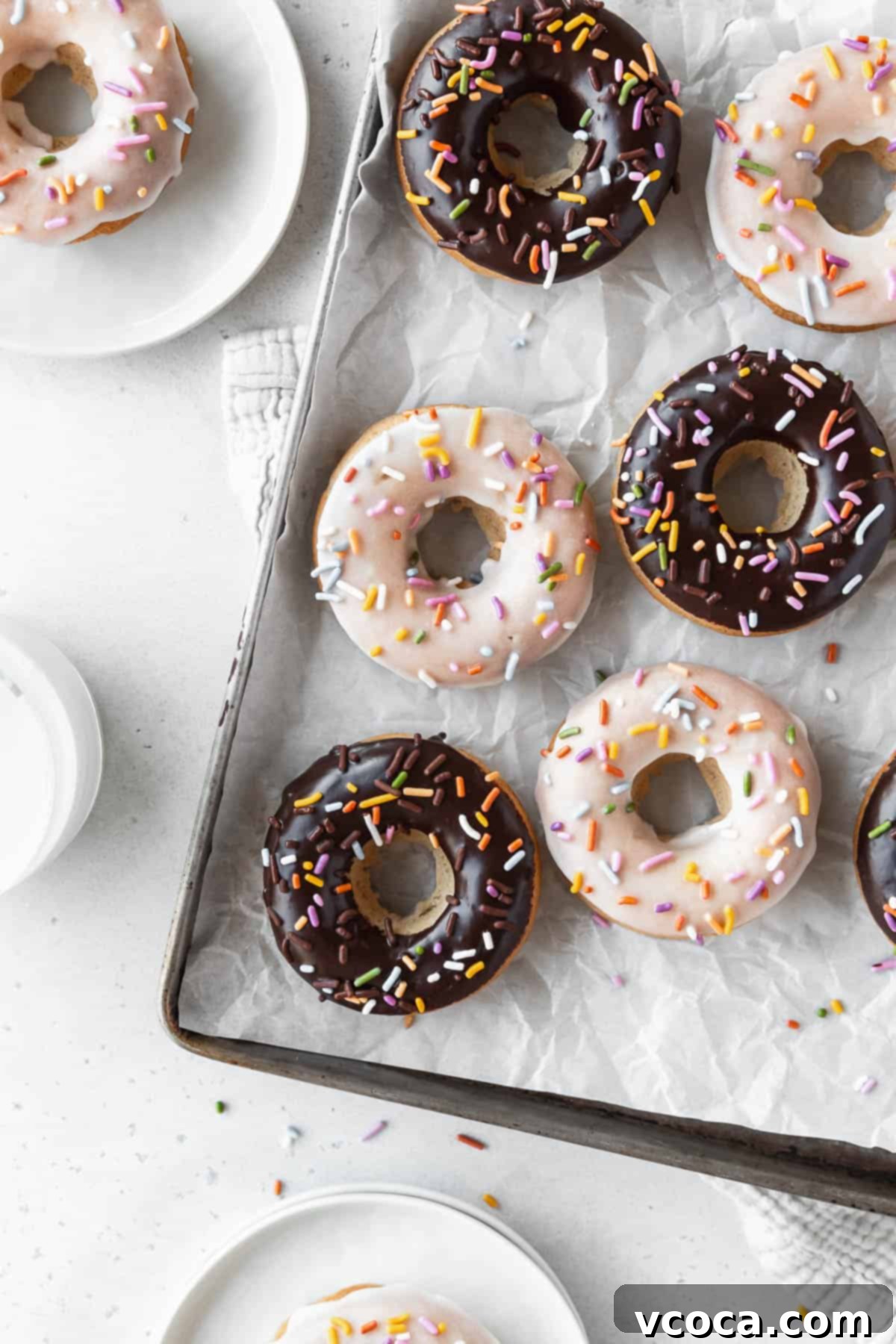 Wholesome Plant-Based Baked Donuts 11 Overhead shot of a parchment lined tray of chocolate and vanilla dipped vegan baked donuts on a white table with small dessert plates holding vanilla glazed vegan doughnuts.