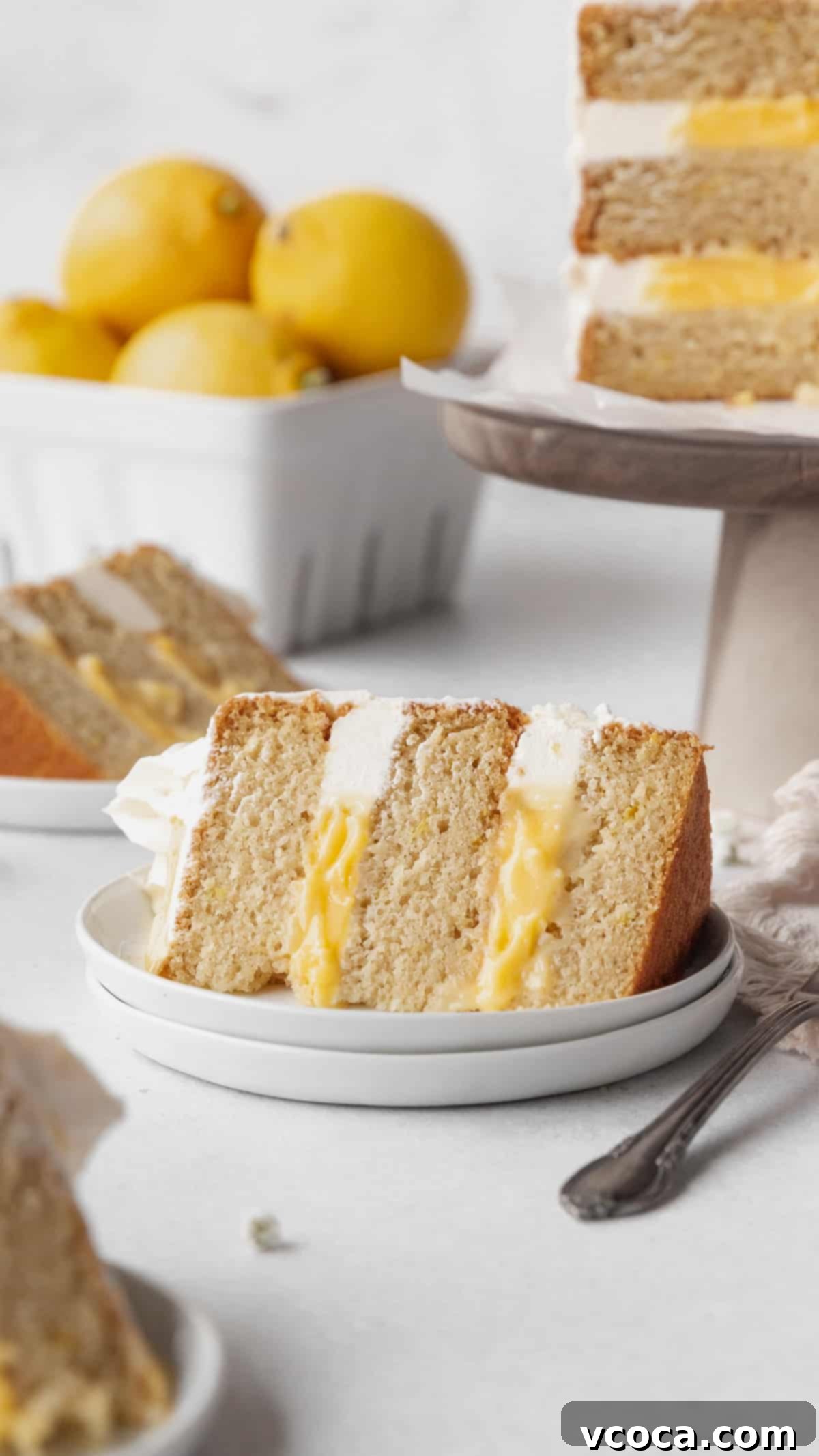 Luscious Lemon Curd Cake 2 A slice of lemon curd cake on a white dessert plate with a cake stand holding the rest of the cake and a ceramic fruit pint holding a handful of lemons in the background.