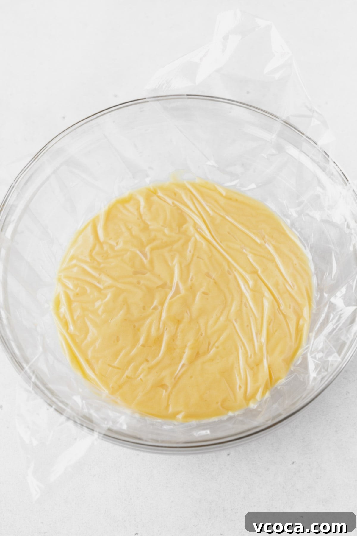 Luscious Lemon Curd Cake 10 A glass bowl of lemon curd with a plastic wrap cover against the surface.