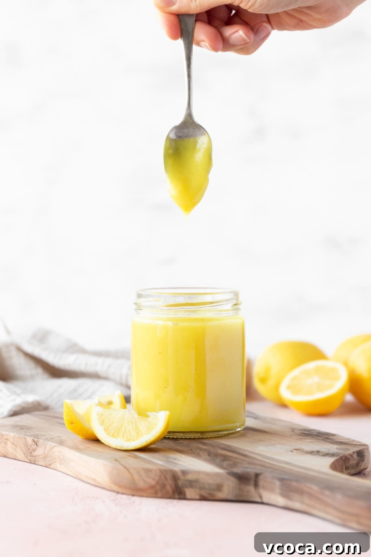 Side on shot of a hand holding a spoon of vegan lemon curd vertically over the full mason jar with the curd slowly dripping off, showcasing its creamy texture.