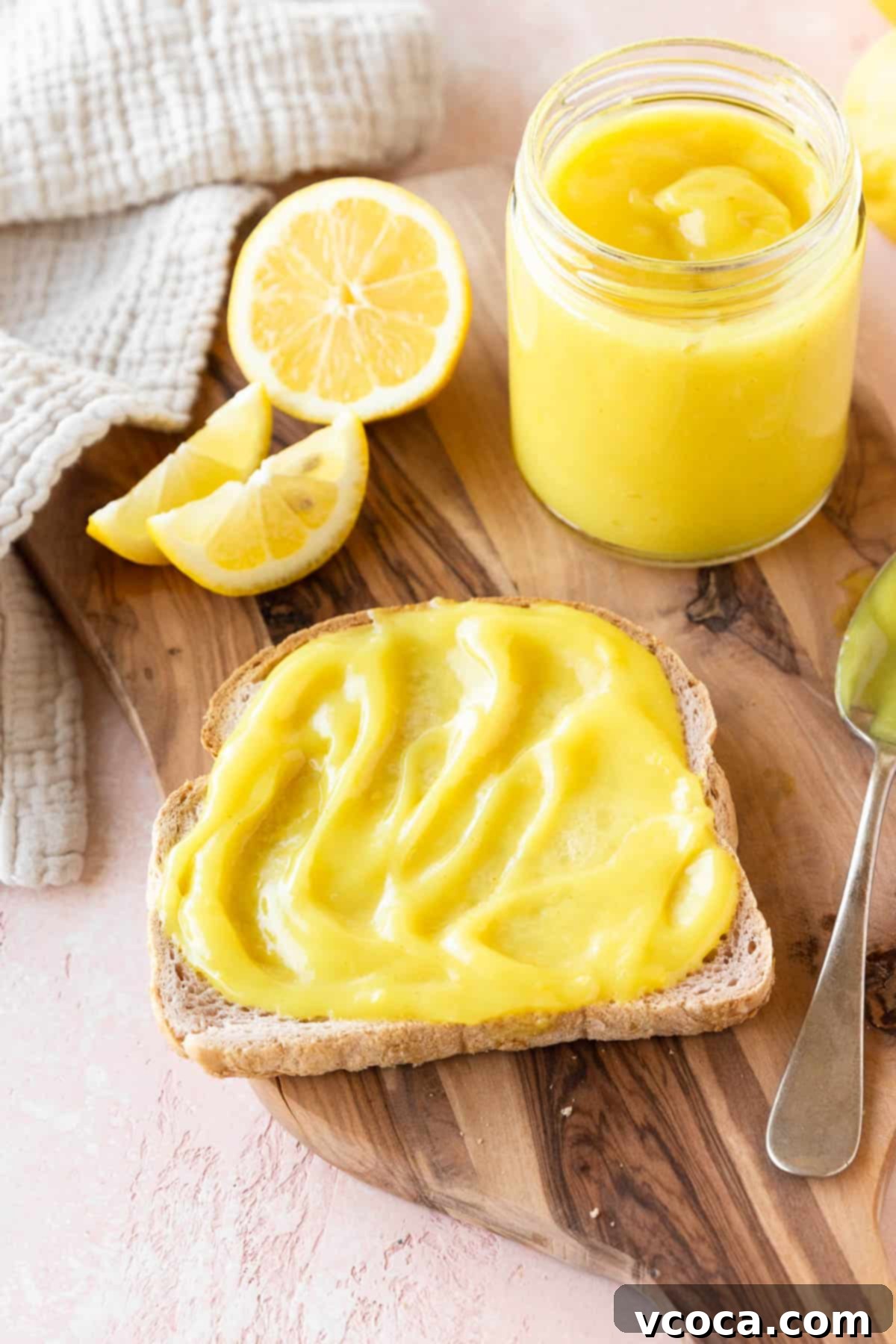 45 degree angle shot of a slice of toast spread with vegan lemon curd on a wooden cutting board with the jar of lemon curd and a halved lemon, ready for breakfast.