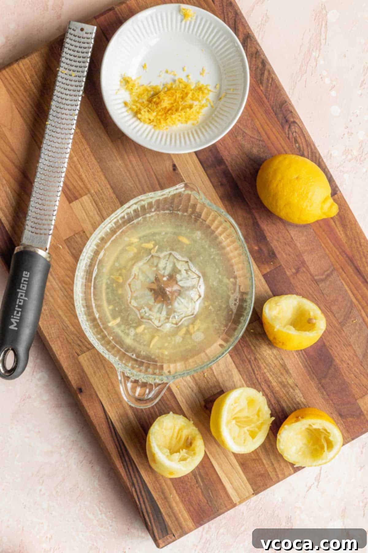 overhead shot of an old fashioned manual lemon juicer bowl filled with juice, a microplane, a plate of lemon zest, and several spent lemons on a wooden cutting board, illustrating the first step of preparation.