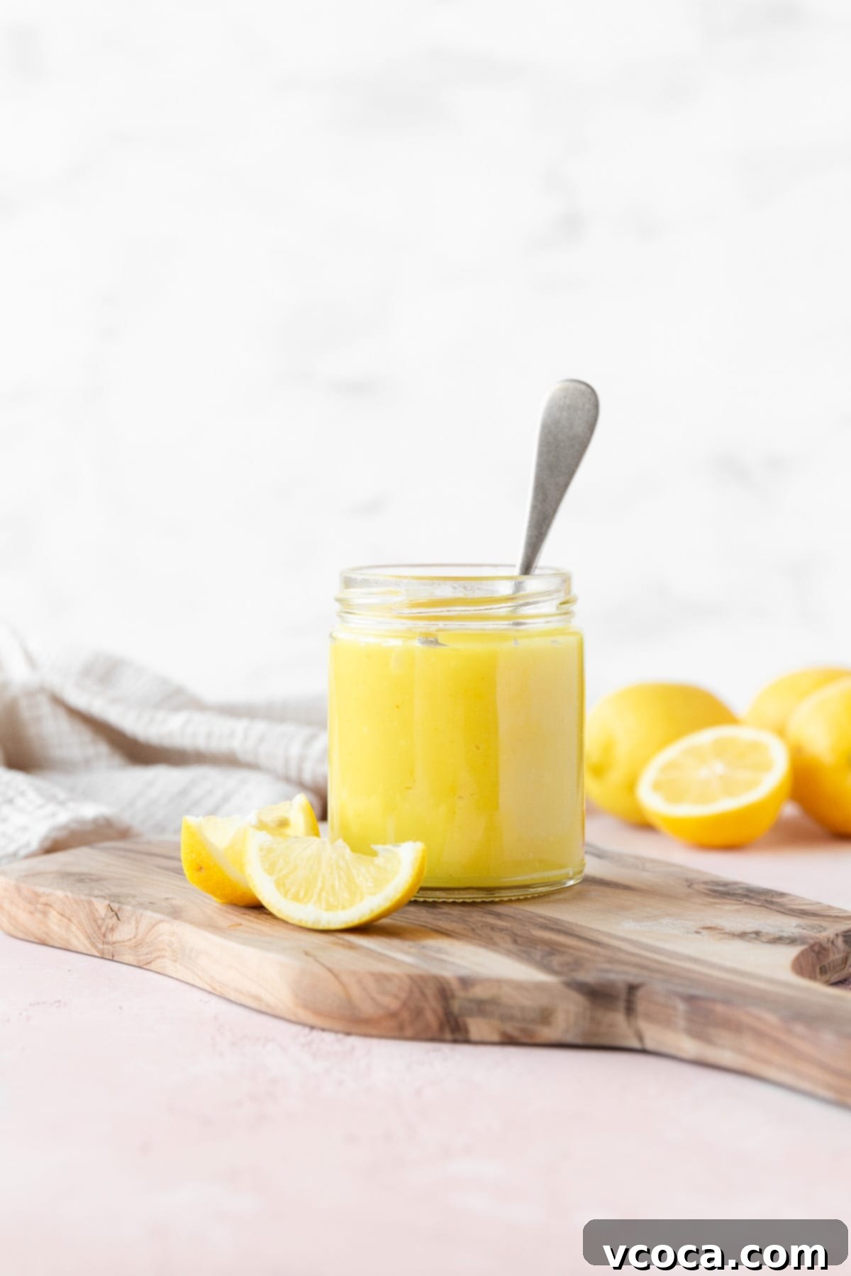 Side on shot of a small mason jar of vegan lemon curd with a silver spoon in it on a wooden cutting board with a few wedges of fresh lemon, ready to be enjoyed.