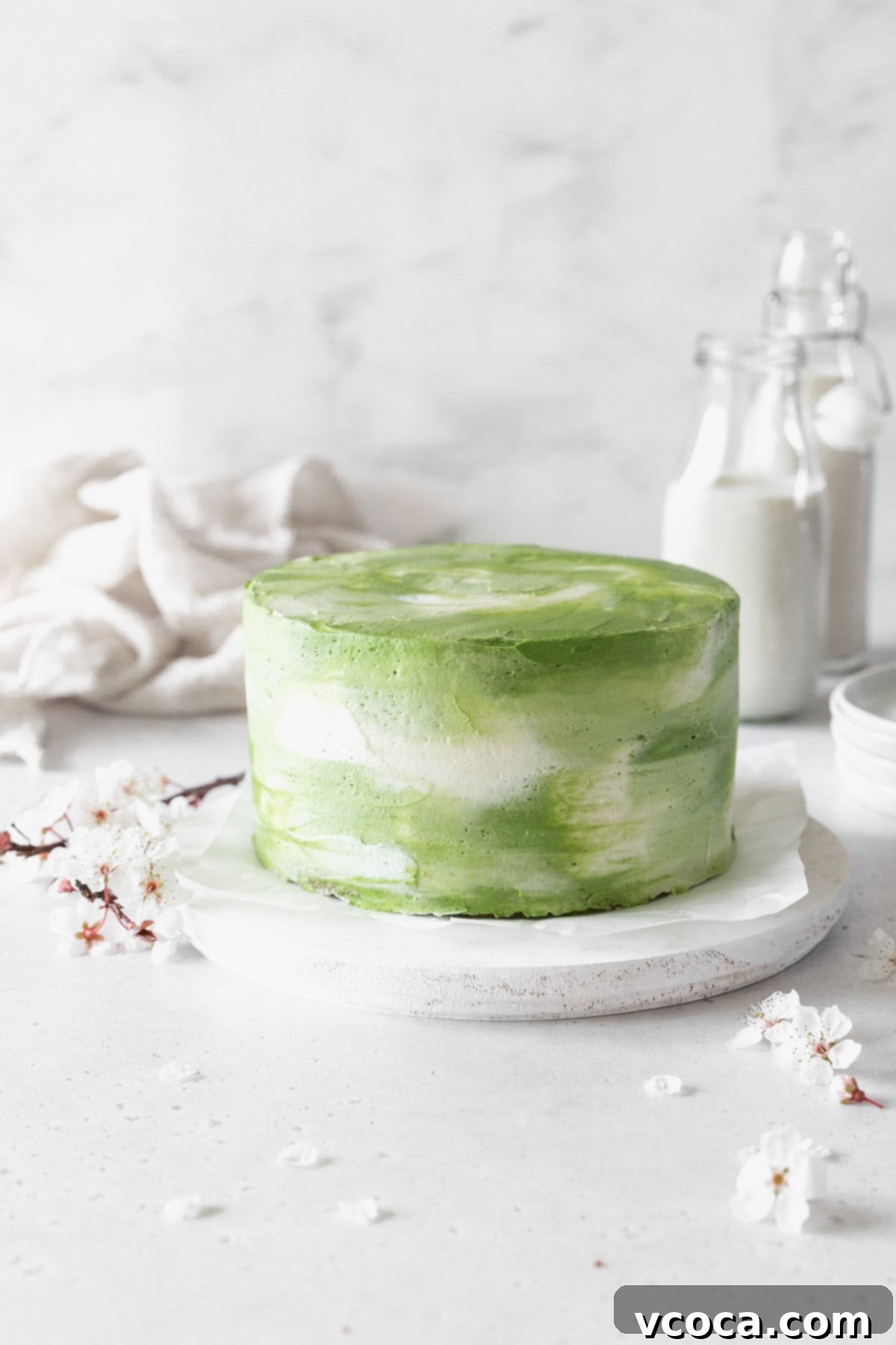 Heavenly Matcha Cake 3 An elegantly iced matcha cake adorned with cherry blossoms on a pristine white cake stand, highlighting its smooth watercolor frosting.