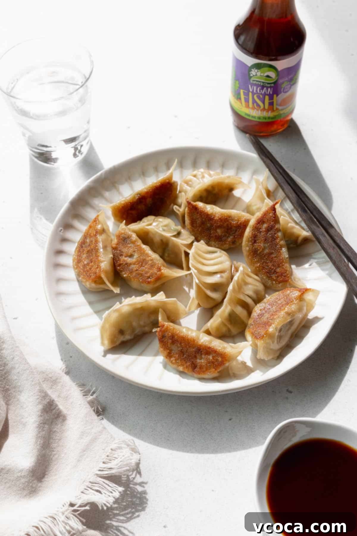 Plant-Based Vegetable Gyoza 4 Angled shot of vegetable gyoza on a white plate with a glass of water and a bottle of vegan fish sauce in the background.