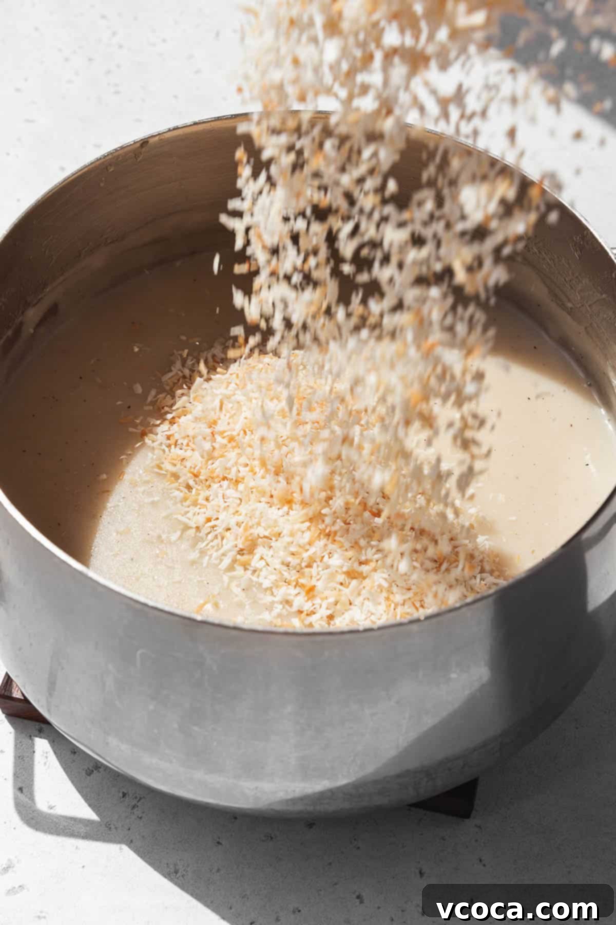 Divine Vegan Coconut Cream Pie Gluten Free 11 Action shot of toasted desiccated coconut being poured into the vegan custard base in the saucepan.