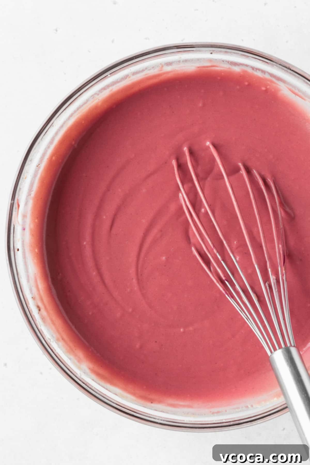 A closeup overhead shot of a glass mixing bowl of red velvet waffle batter with a whisk.