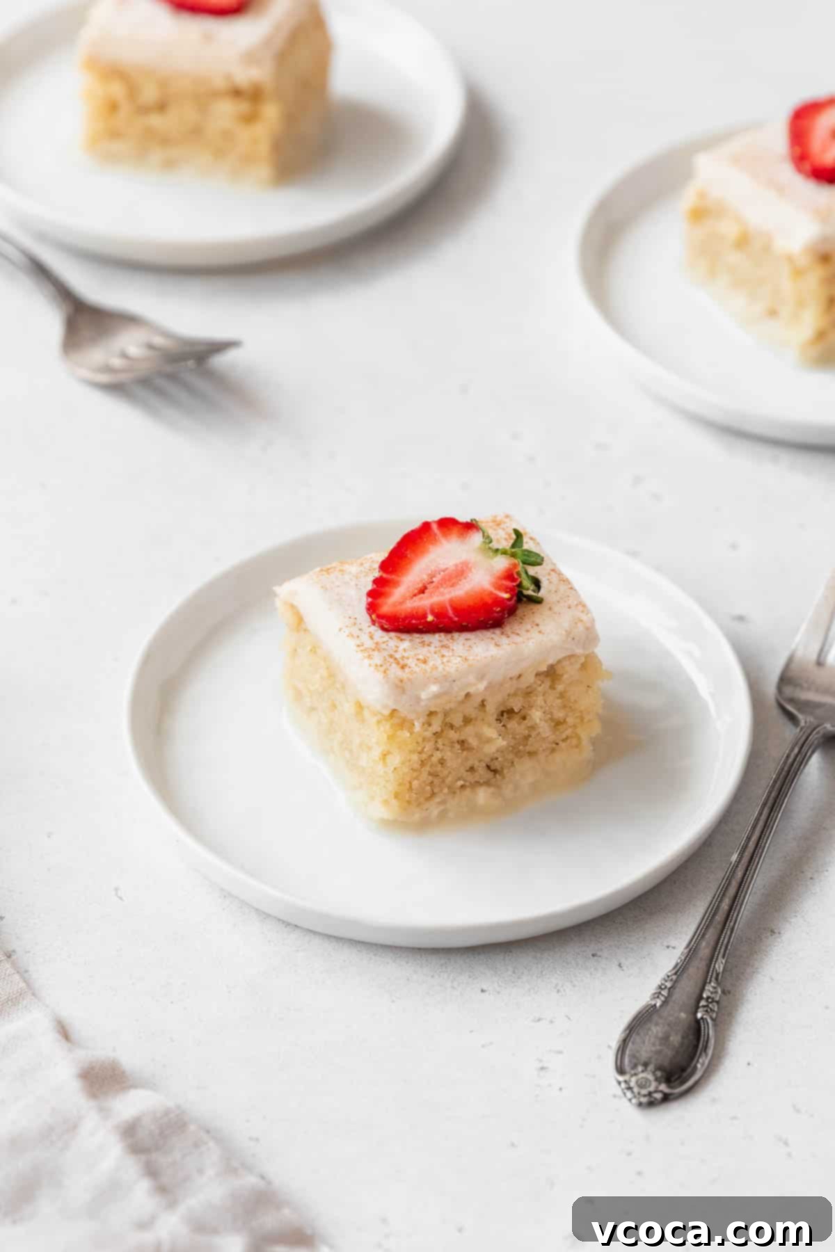 Three generous slices of gluten-free and dairy-free tres leches cake, elegantly presented on white dessert plates, each garnished with vibrant red strawberries. The cake itself boasts a rich, creamy topping and a visibly moist texture, inviting a delicious indulgence.