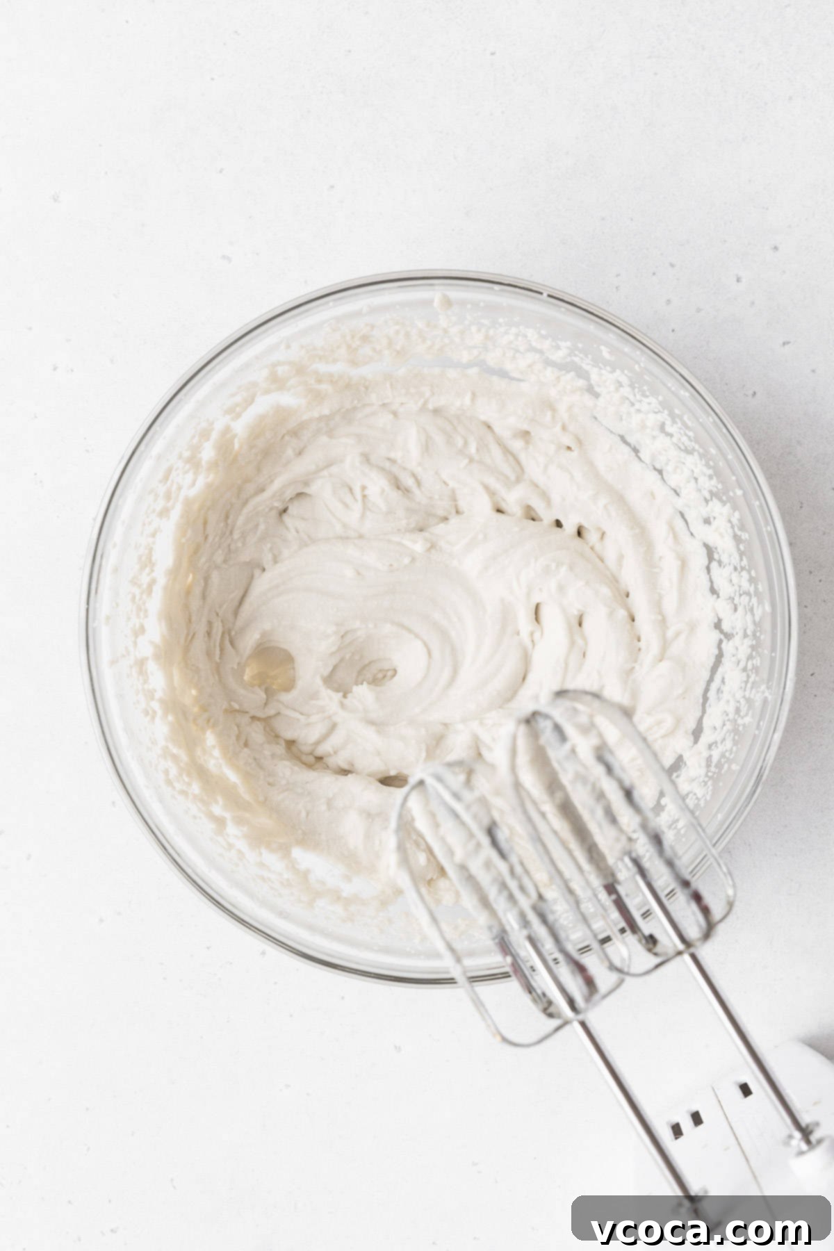 The process of whipping fluffy non-dairy cream in a clear glass mixing bowl, using a hand mixer to achieve the perfect light and airy texture for the cake topping.