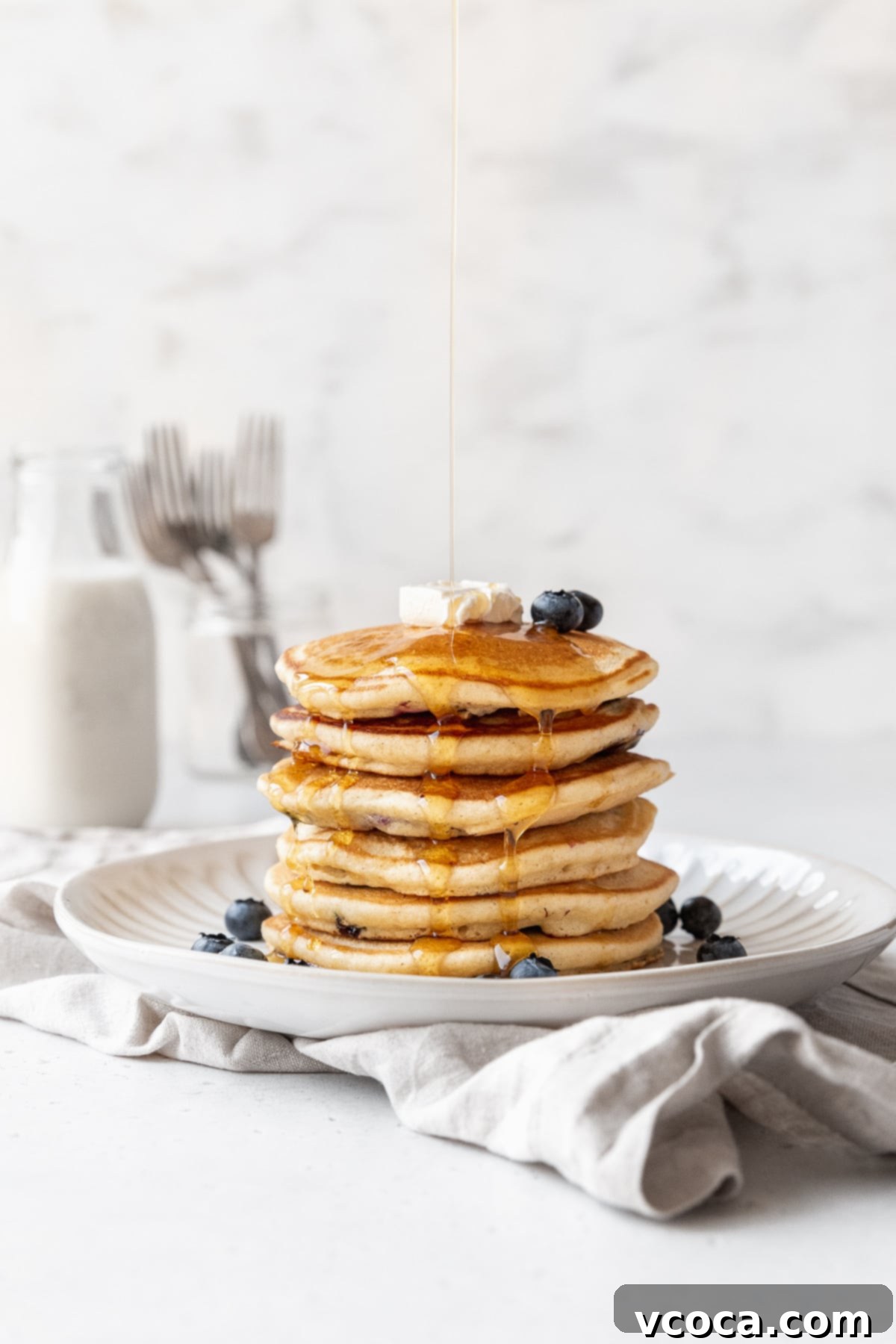 Distance side-on action shot of syrup being drizzled on a shortstack of blueberry studded muffin mix pancakes.