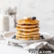 action shot of syrup being poured onto a stack of blueberry muffin mix pancakes topped with a pad of butter and extra fresh blueberries.