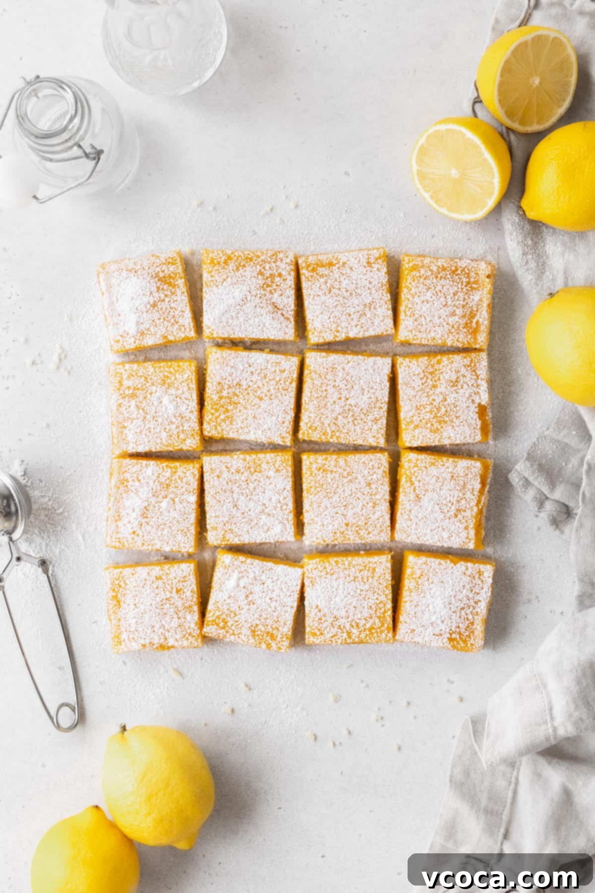 Bright and Zesty Dairy Free Lemon Bars 20 A pan of dairy free lemon bars cut into squares on the counter, with halved lemons and powdered sugar nearby.