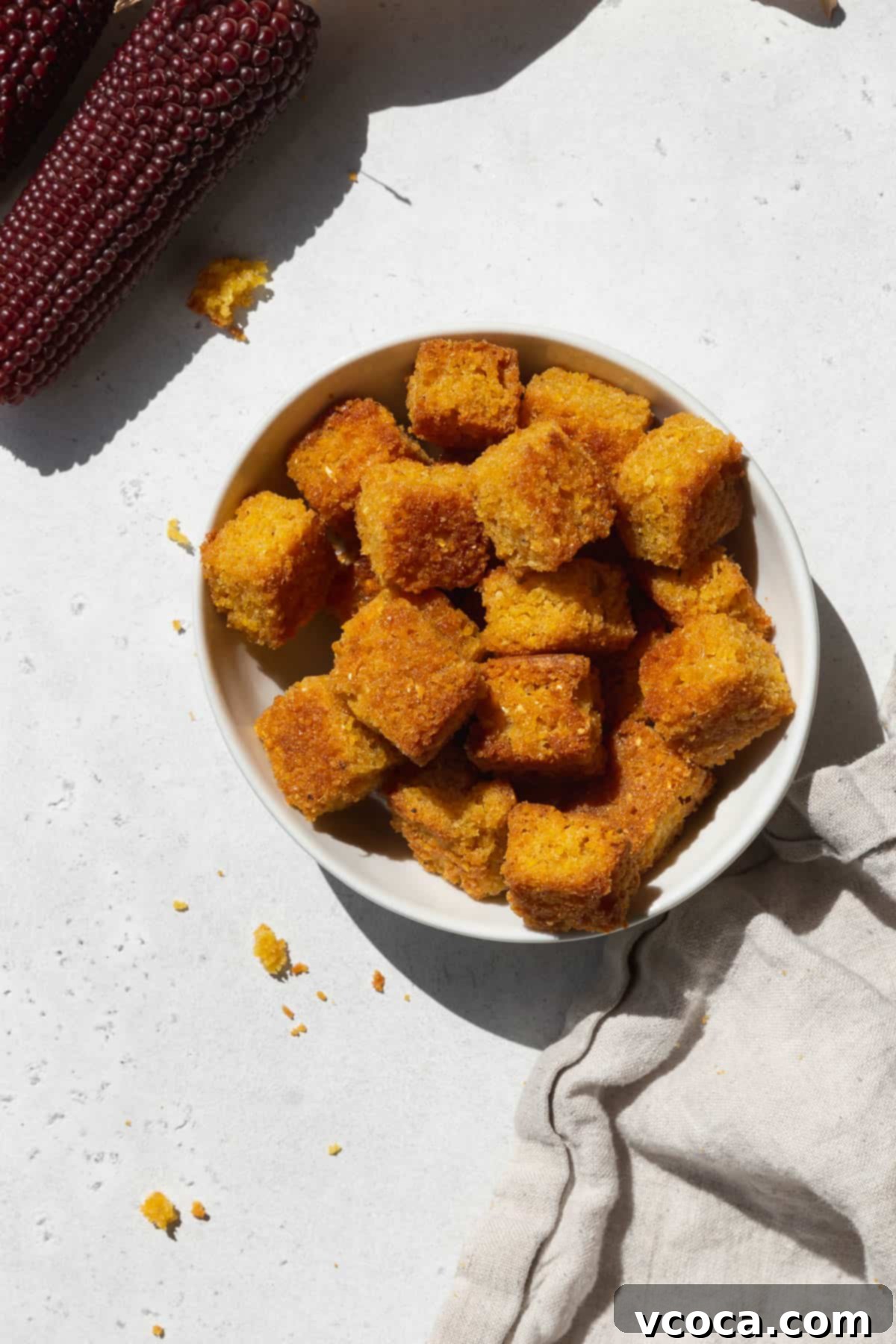 An overhead shot of a small bowl of cornbread croutons with a napkin and a stalk of heirloom corn.