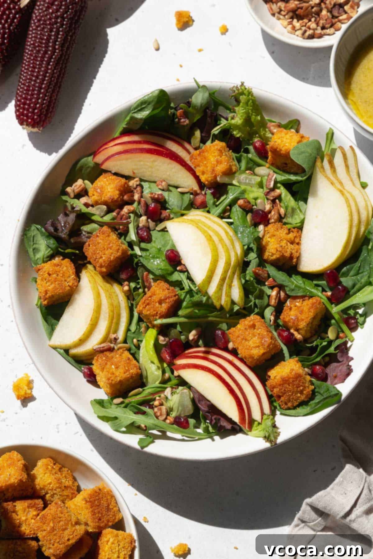 An overhead shot of a large white bowl of salad with cornbread croutons, sliced pears and apples, and pomegranate seeds.