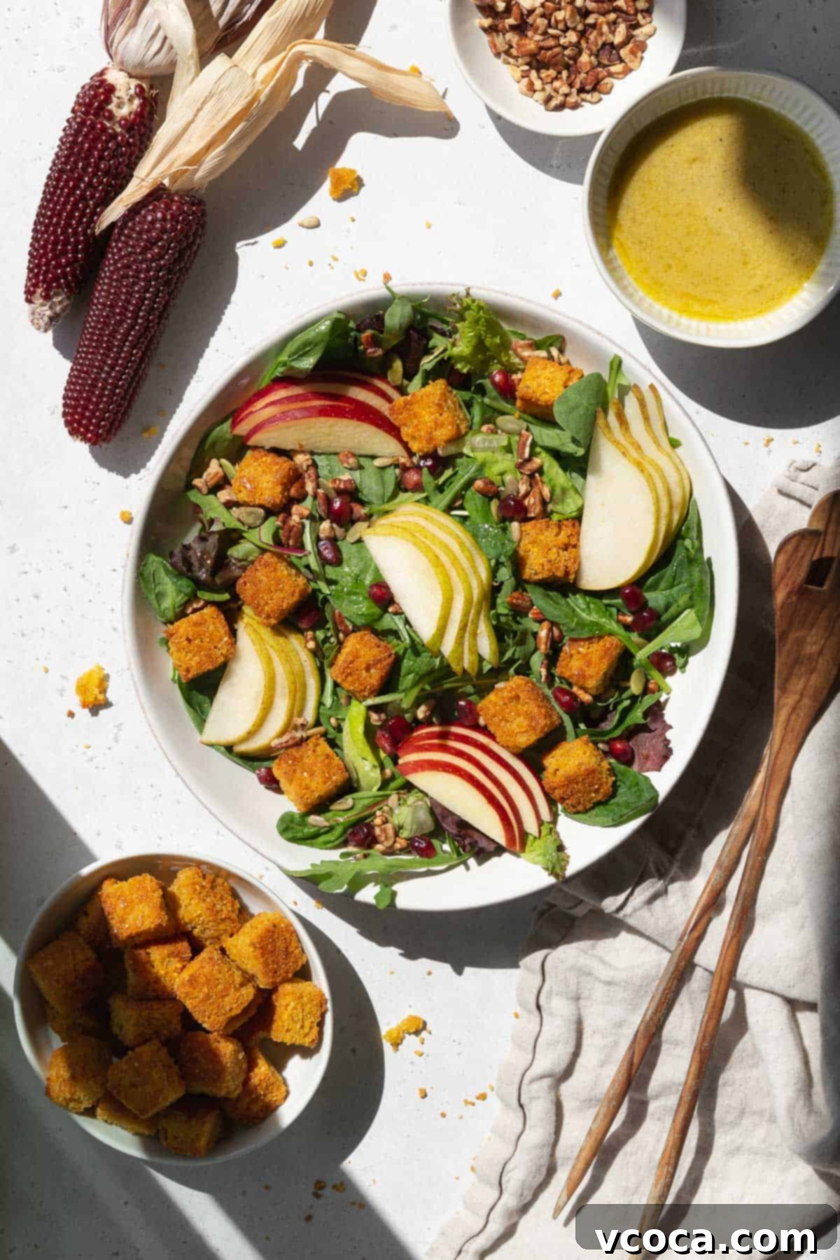 A spread of salad with cornbread croutons, pears, apples, dressing, and salad tongs.