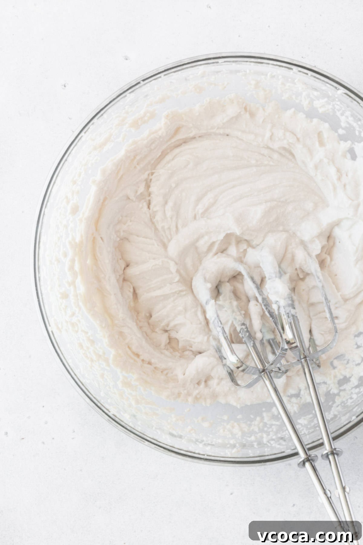 A bowl of creamy vegan whipped cream, with two hand mixer beaters resting inside, indicating readiness for the next step of preparation.