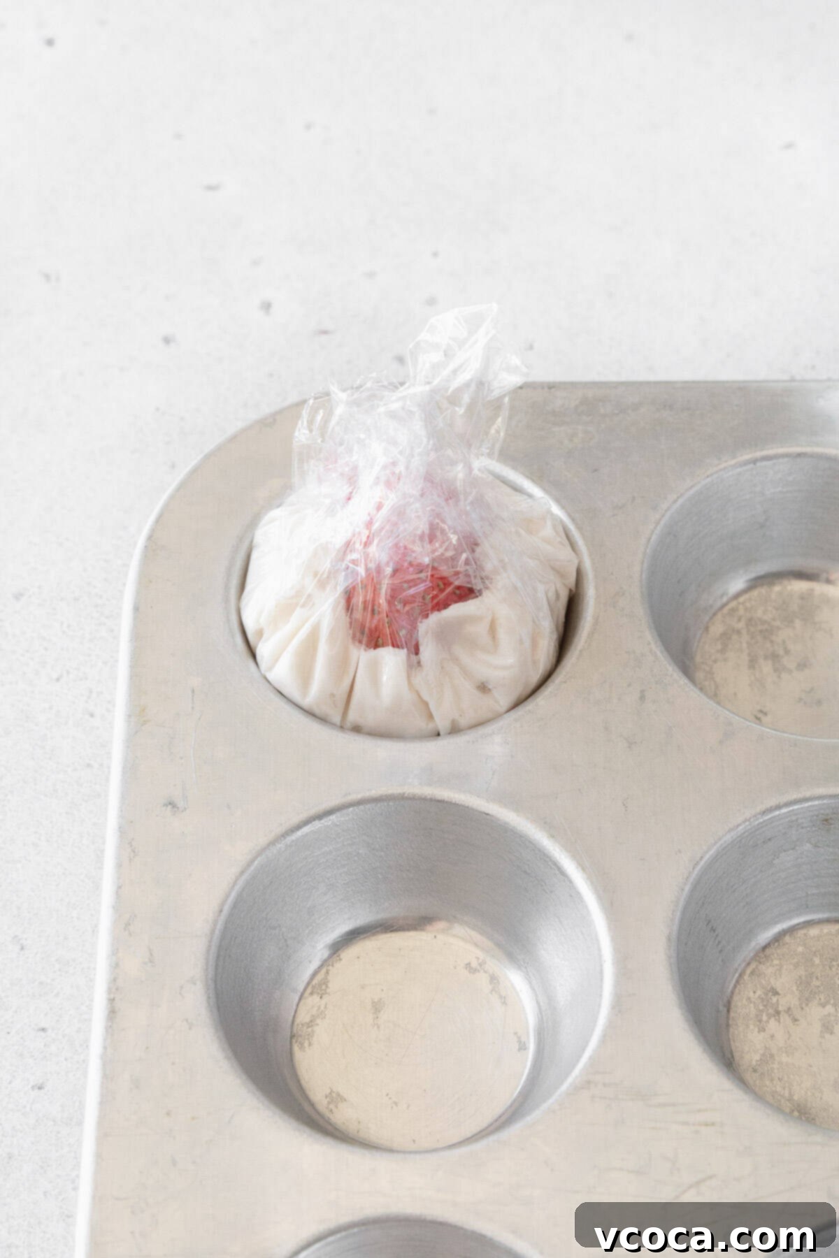 Plastic wrap expertly twisted over a strawberry and whipped cream portion within a mini muffin tin, sealing the filling for chilling.