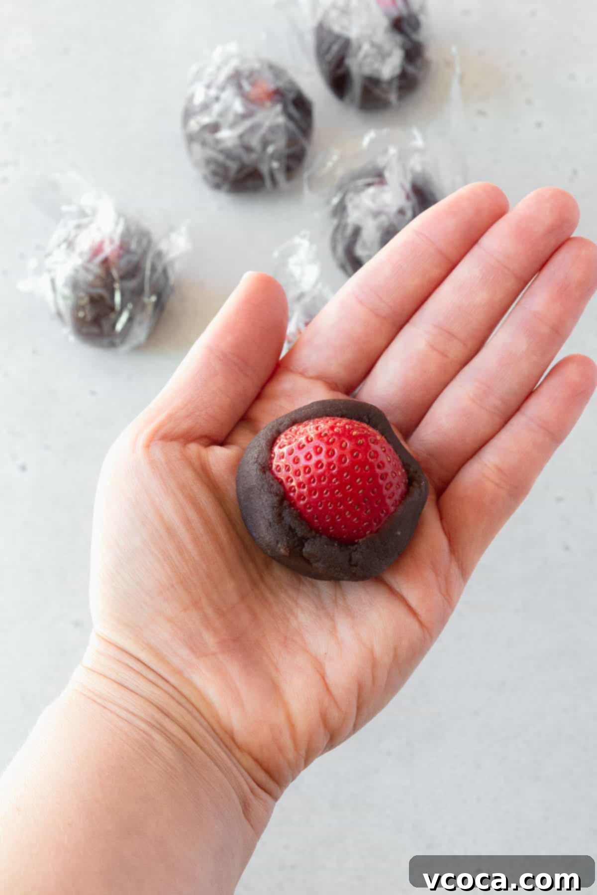 A hand carefully holds a fresh strawberry, half-wrapped in a layer of smooth, sweetened red bean paste, illustrating the first stage of the filling process.