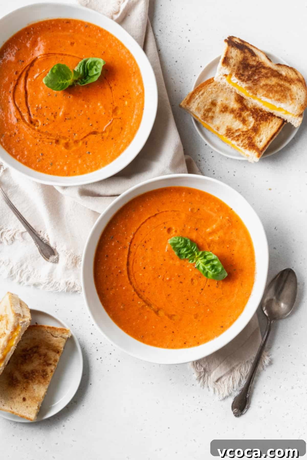 Heartwarming Roasted Tomato Soup 11 Two cozy bowls of creamy roasted tomato soup, beautifully garnished with fresh basil, placed alongside small plates of perfectly grilled cheese halves and spoons on a pristine white surface.