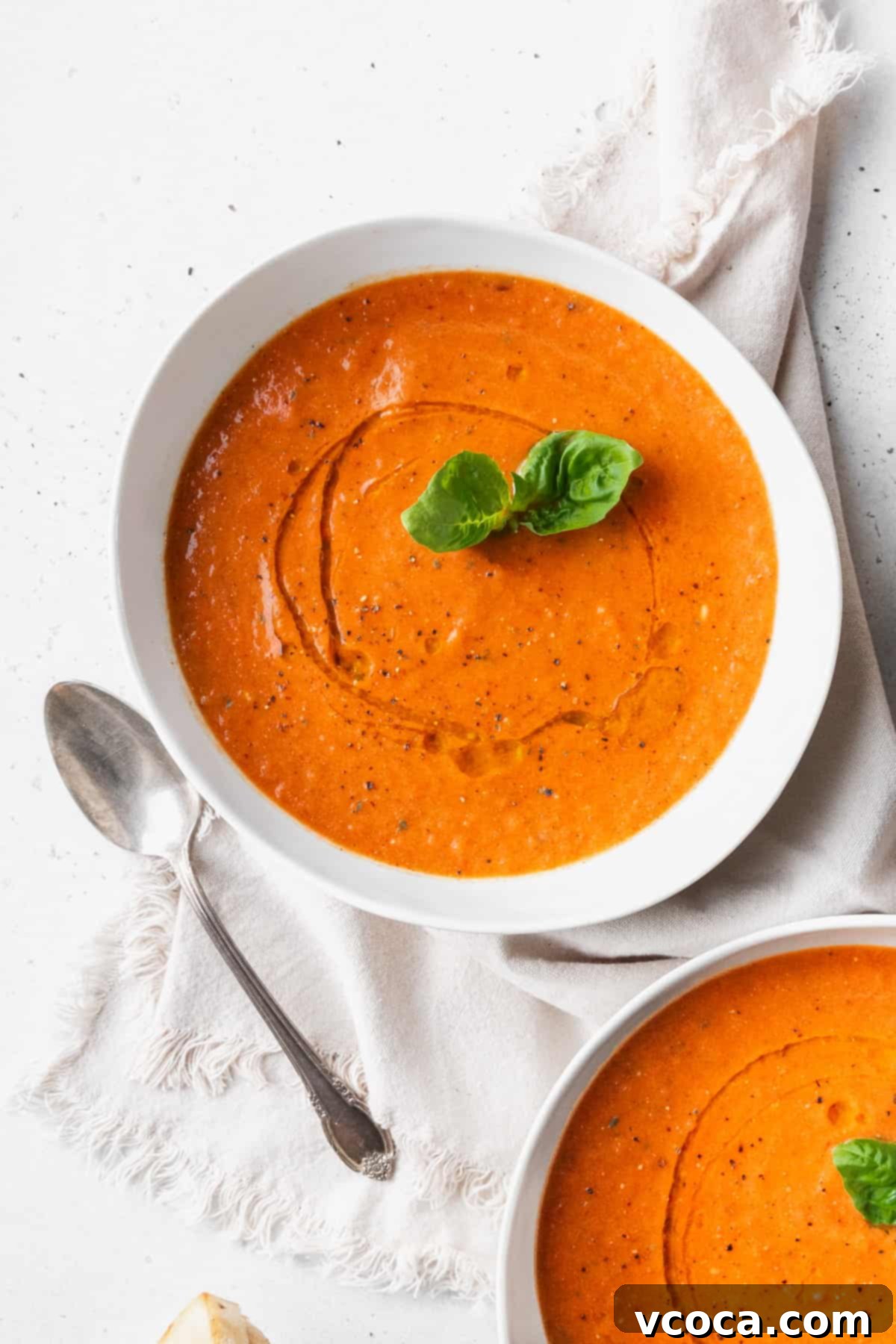 Heartwarming Roasted Tomato Soup 10 A beautifully presented white bowl of creamy roasted tomato and basil soup, garnished with a fresh sprig of basil, placed on a light linen next to a silver spoon.