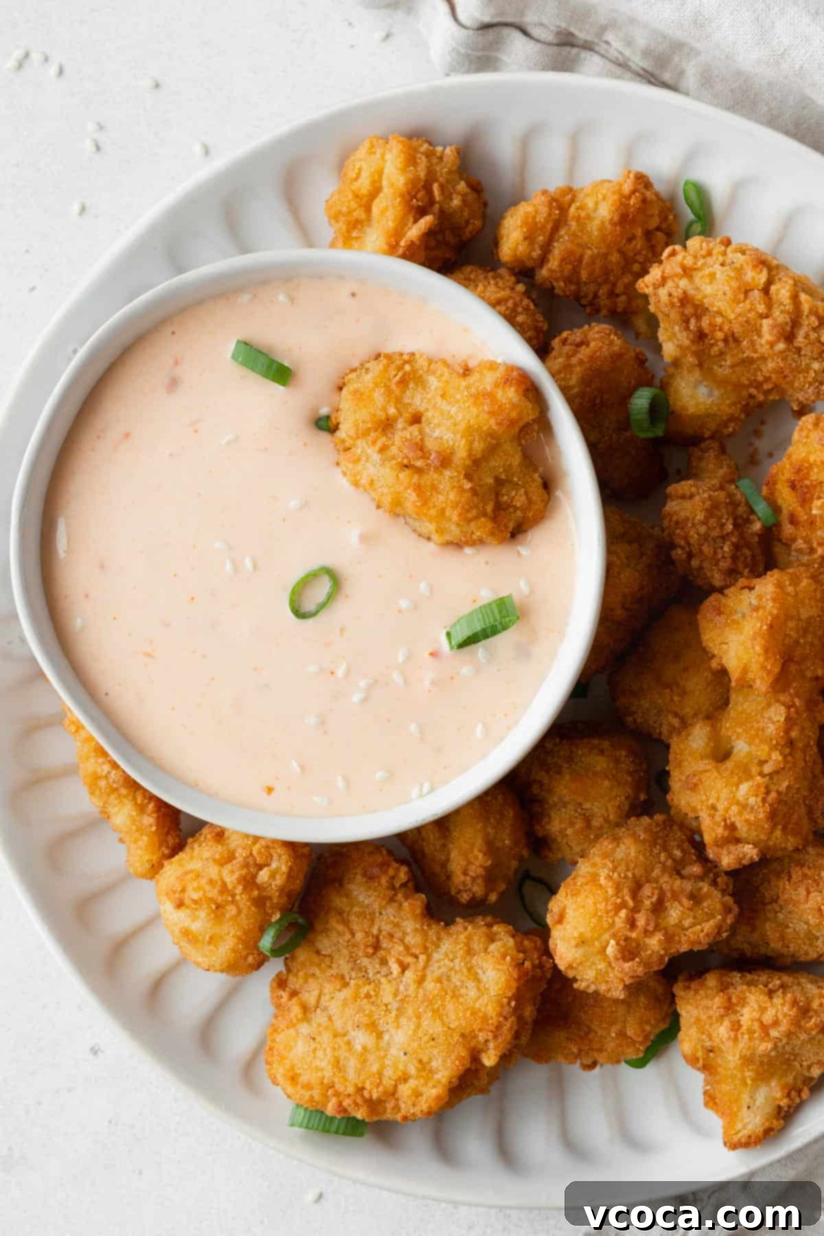 A plate of golden cauliflower bites generously drizzled with a rich, creamy Bang Bang sauce, with a small bowl of the sauce beside it.