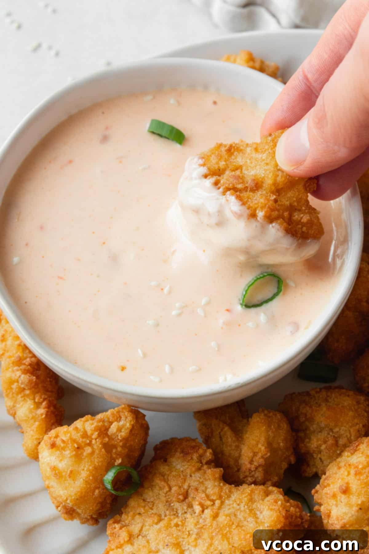 A hand holding a deep-fried cauliflower floret, dipping it into a bowl of creamy, easy homemade Bang Bang Sauce, showcasing its perfect consistency.