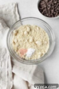 Dry ingredients for cookies, including almond flour and salt, measured into a clear glass bowl, ready for mixing.