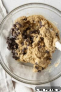 Plenty of chocolate chips being folded into creamy almond flour cookie dough in a large glass bowl with a spatula.