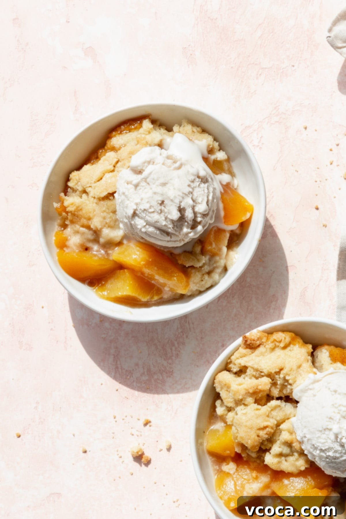 Homemade Peach Cobbler 3 A bowl of peach cobbler with canned peaches and a scoop of vanilla ice cream on a blush colored surface.