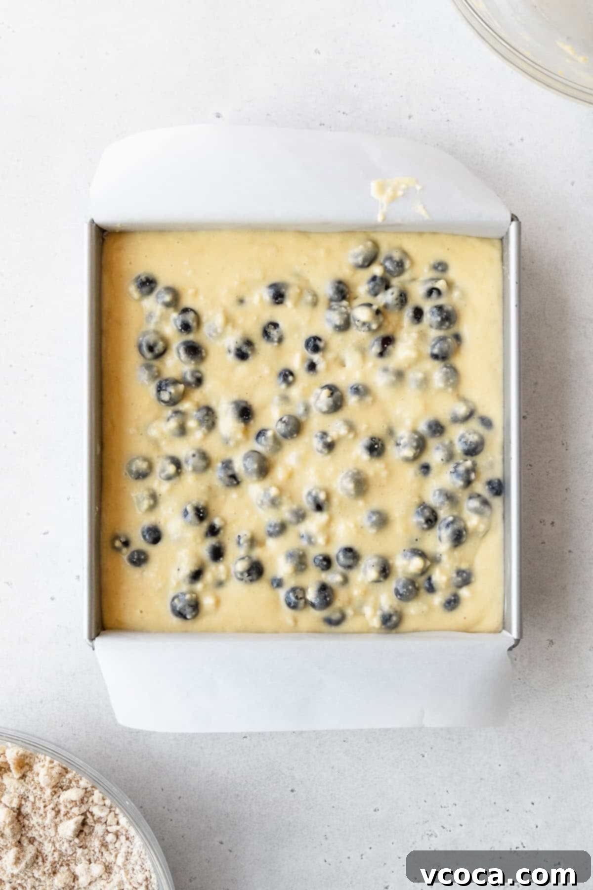 Blueberry Coffee Cake 14 Blueberry cake batter layered in a square pan on a white surface, ready for the crumb topping.
