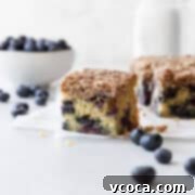 Blueberry Coffee Cake 19 A slice of fluffy blueberry coffee cake with a generous streusel topping, served on a white surface with scattered fresh blueberries.