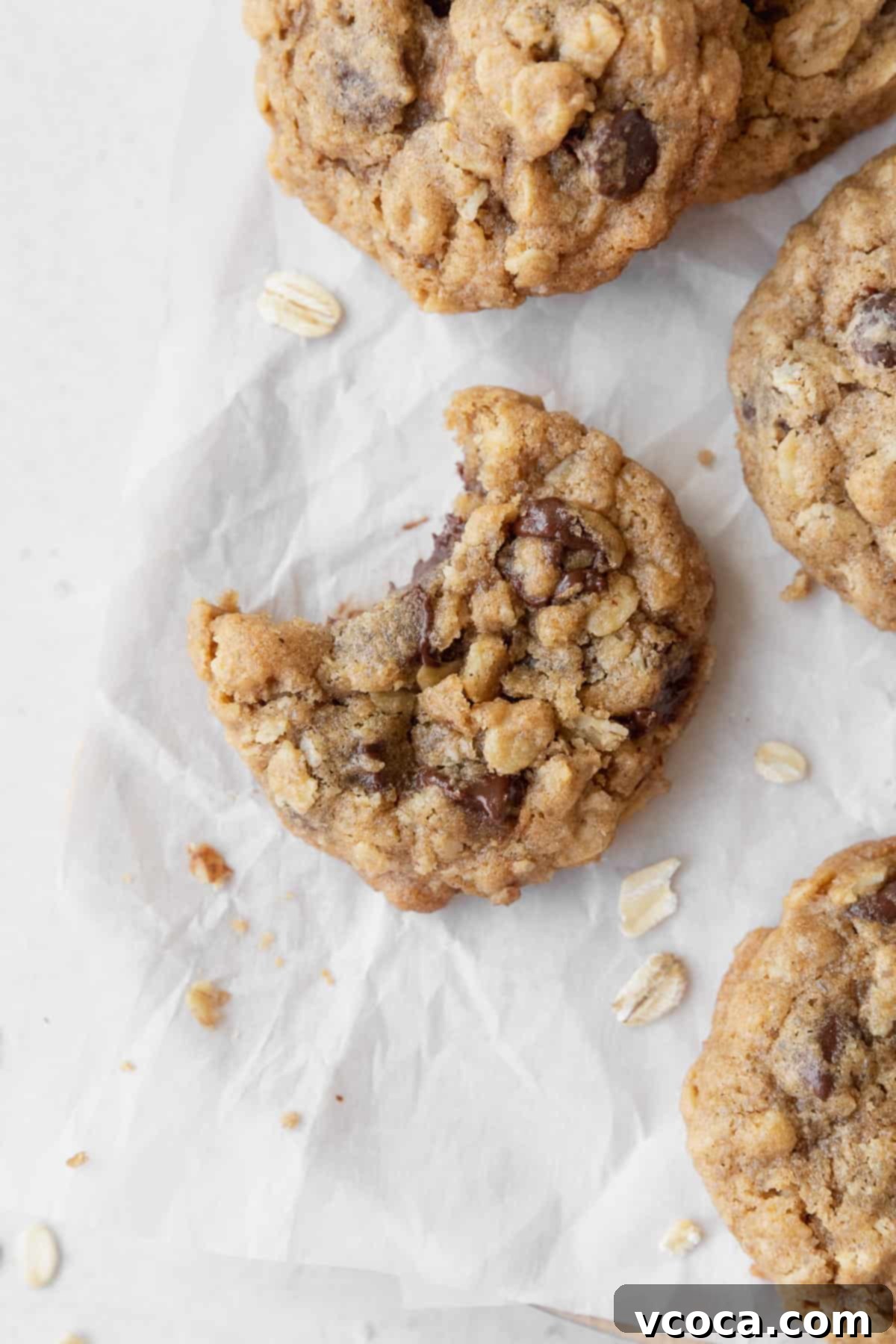 Delicious Gluten-Free Oatmeal Cookies 2 A perfectly baked, slightly bitten gluten-free oatmeal cookie, showcasing its chewy interior with visible oats and chocolate chips. It rests on parchment paper, surrounded by more cookies and scattered rolled oats.