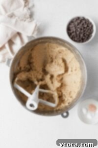 The wet ingredients for gluten-free oatmeal cookies, including creamed butter, sugars, egg, and vanilla, in a metallic mixing bowl, with a small bowl of chocolate chips nearby.