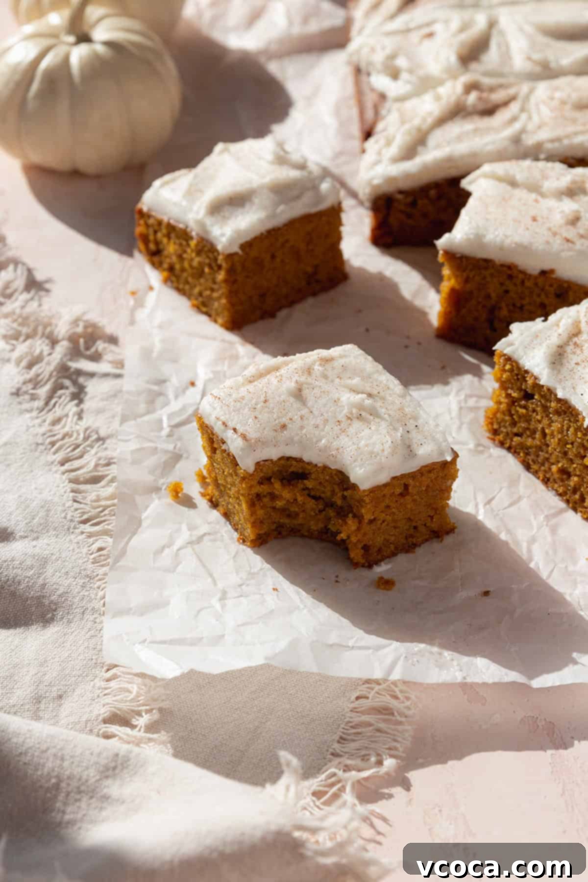 Spiced Pumpkin Squares 12 A perfectly bitten square of homemade pumpkin bar with cream cheese frosting, on parchment paper with more squares ready to be shared.