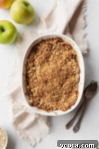 Baked apple crisp on a beige linen with a spoon, fork, and some green apples placed next to it.