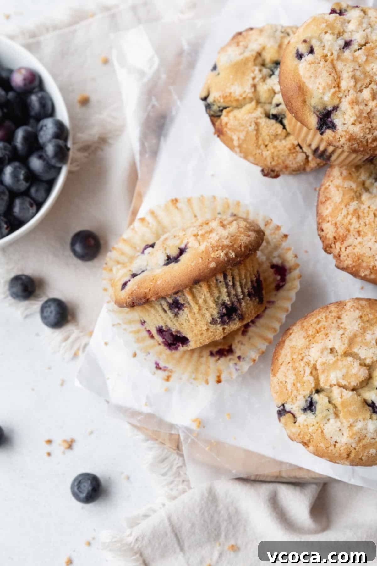 Guilt-Free Blueberry Muffins 11 A blueberry muffin laying on its side on a paper liner, with more muffins on the right and a bowl of blueberries on the left.
