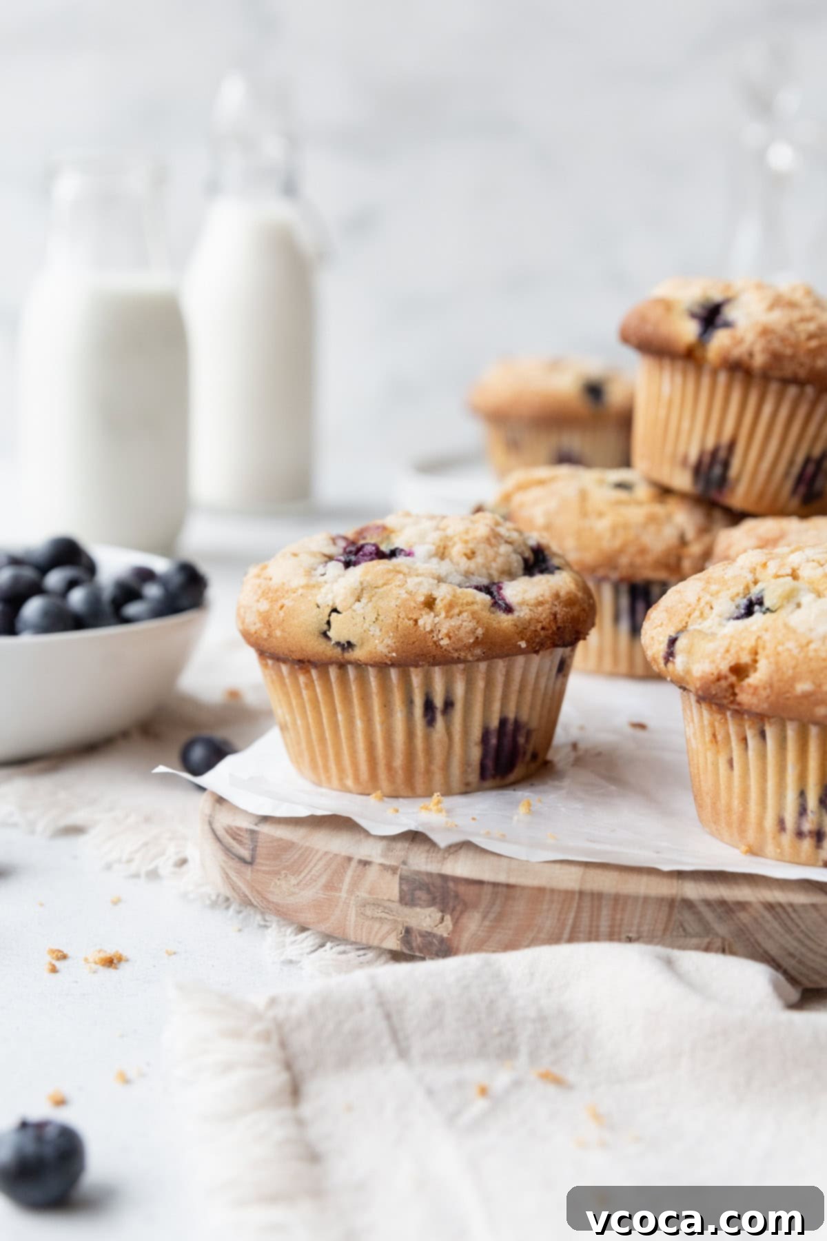 Guilt-Free Blueberry Muffins 3 Gluten-free blueberry muffins on a wooden board lined with parchment paper, with a bowl of blueberries on the side and milk jars in the background.