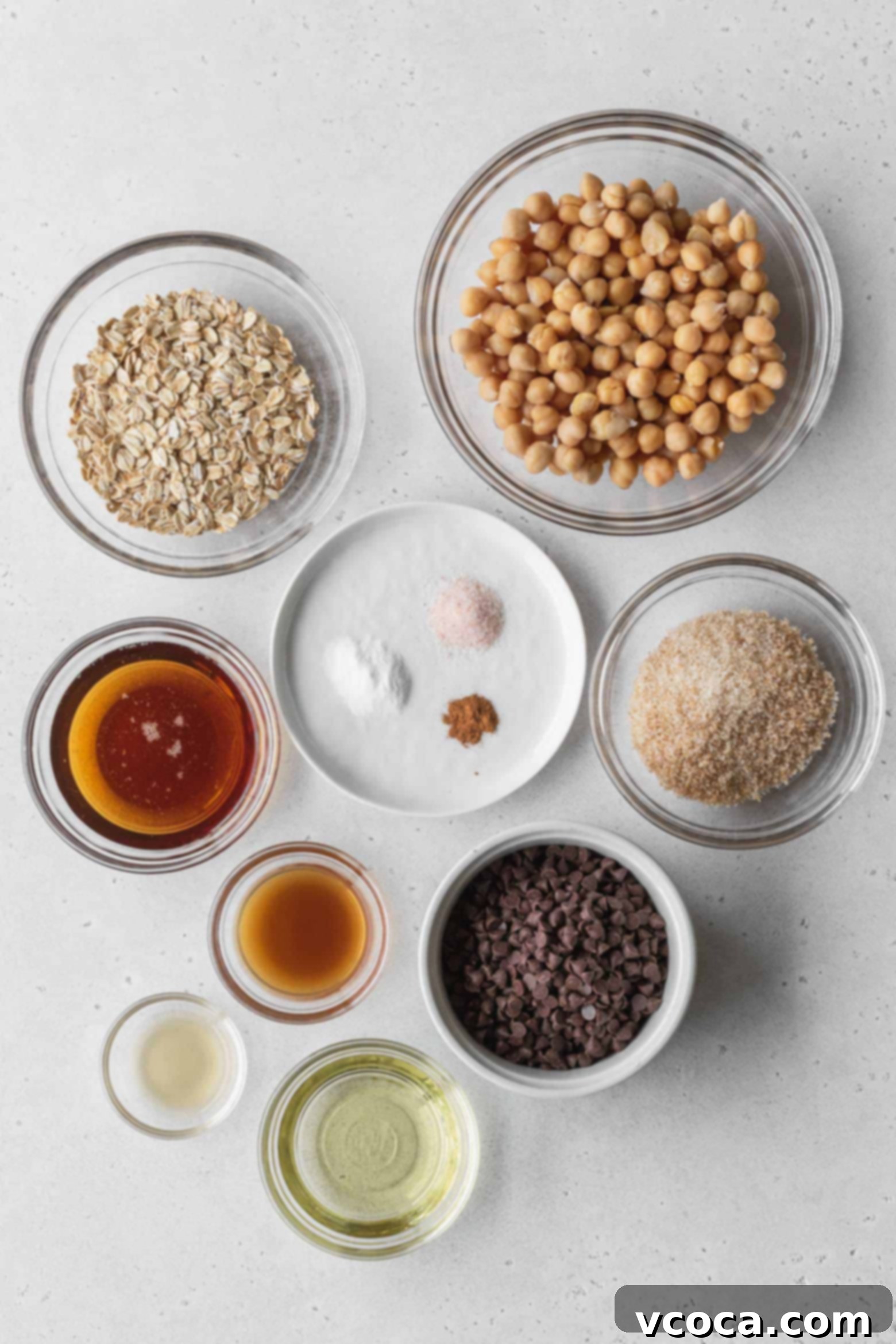 Various ingredients for chocolate chip chickpea cookies, including chickpeas, oats, sugar, and chocolate chips, carefully measured into separate bowls on a wooden surface.