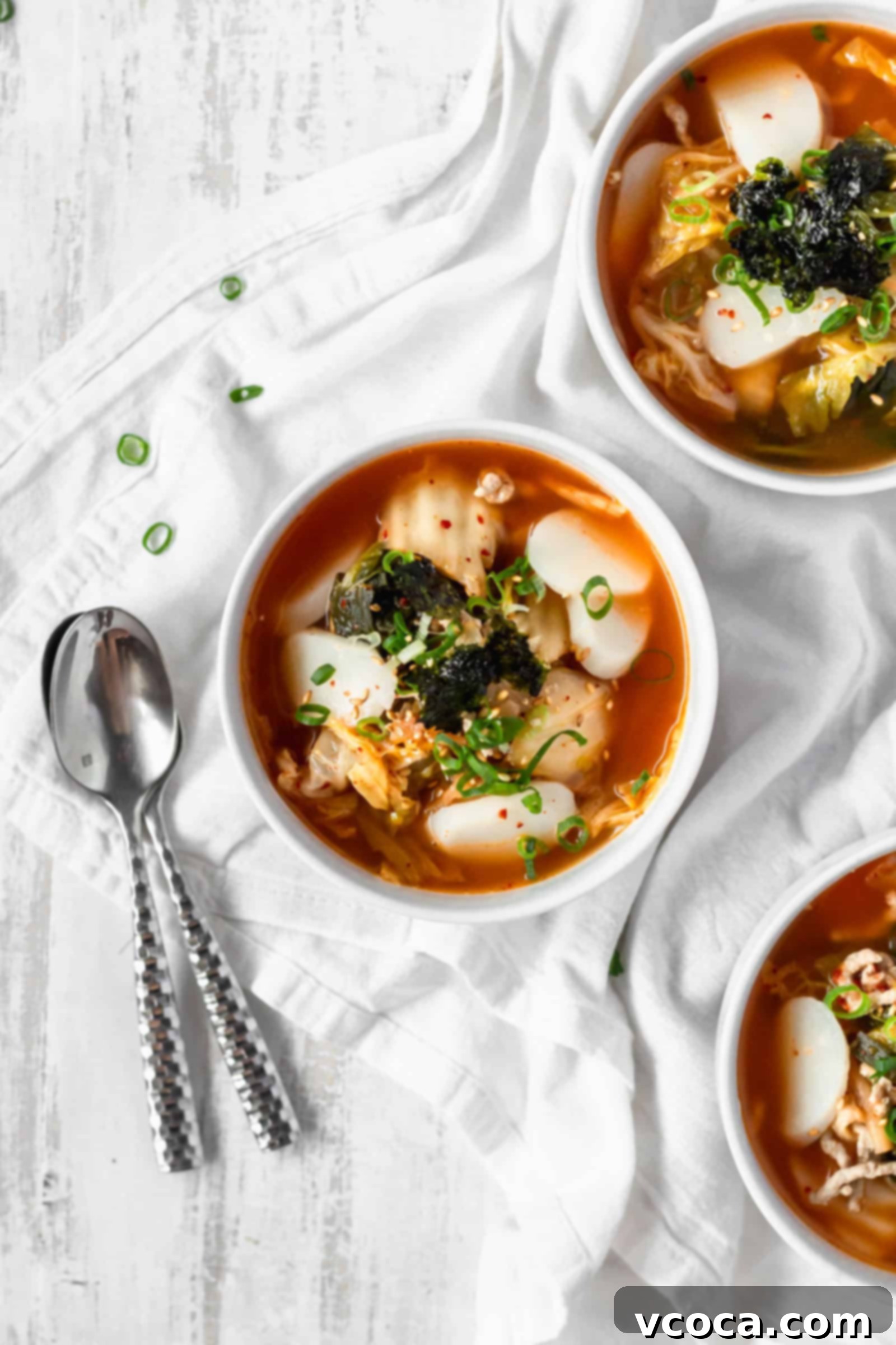 Spicy Kimchi Rice Cake Soup 2 Three bowls of tteokguk on a white surface with two silver spoons. Each bowl is beautifully garnished, showcasing the chewy rice cakes and savory broth.