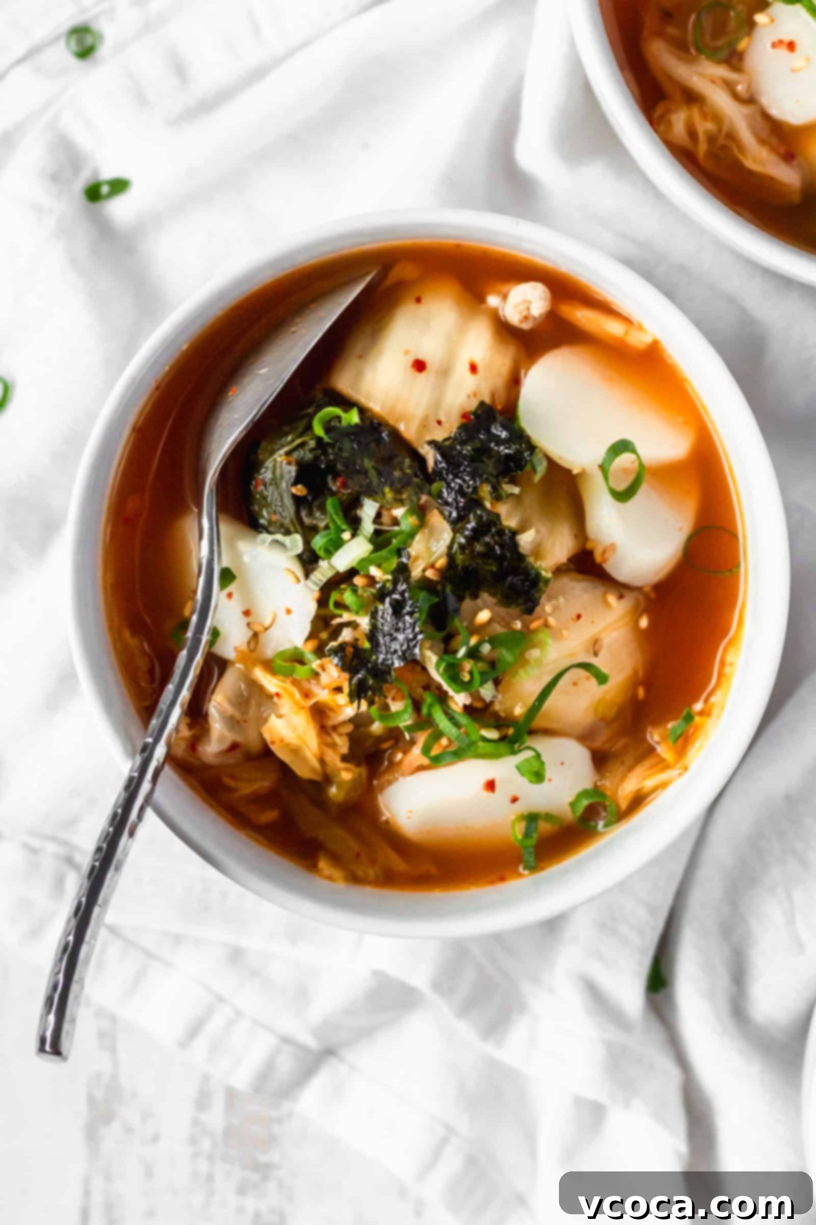 Spicy Kimchi Rice Cake Soup 3 An overhead shot of a bowl of Korean rice cake soup on a white surface, garnished with seaweed and sesame seeds.