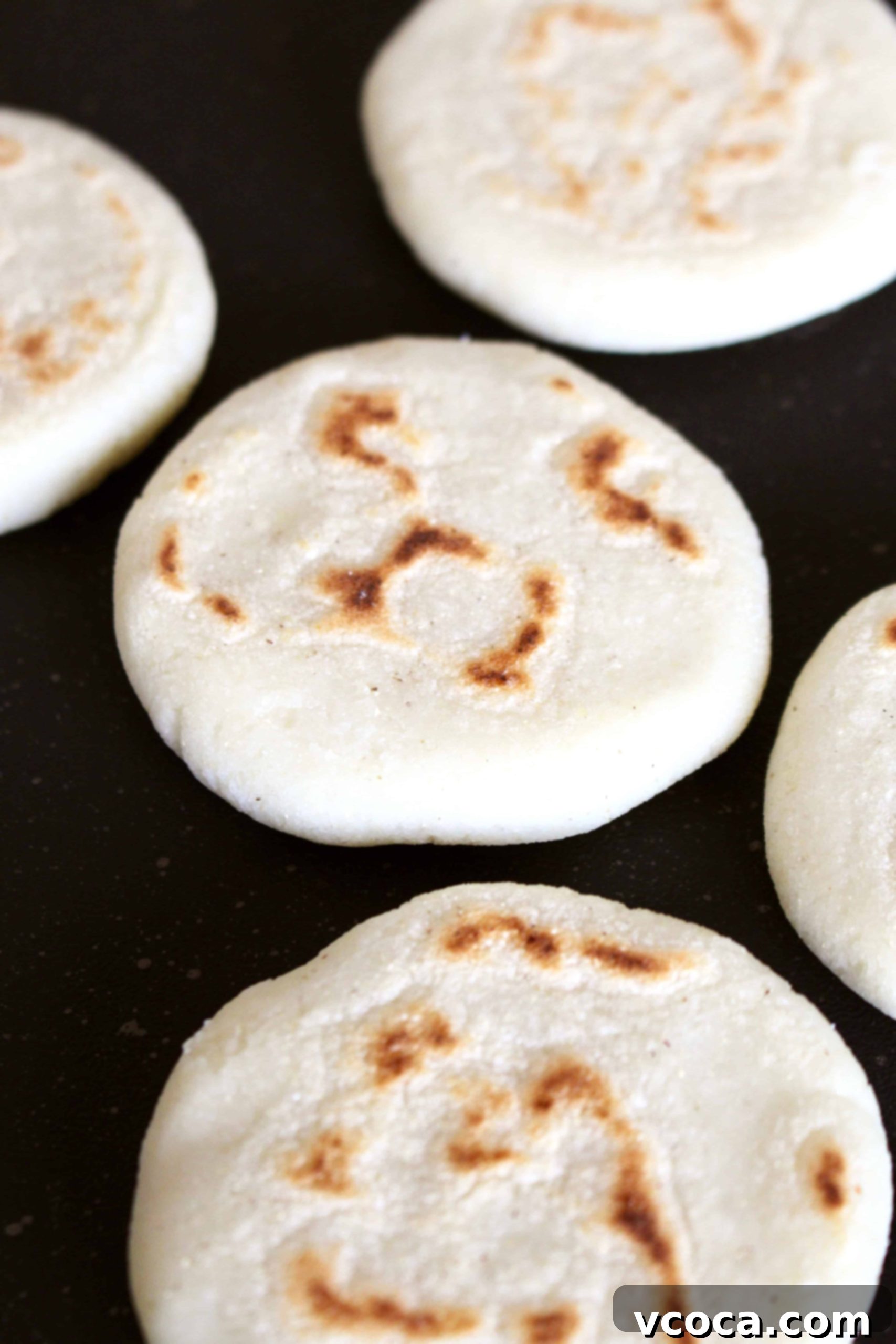 Several arepas cooking on a dark metal pan, showing their developing golden-brown crusts.