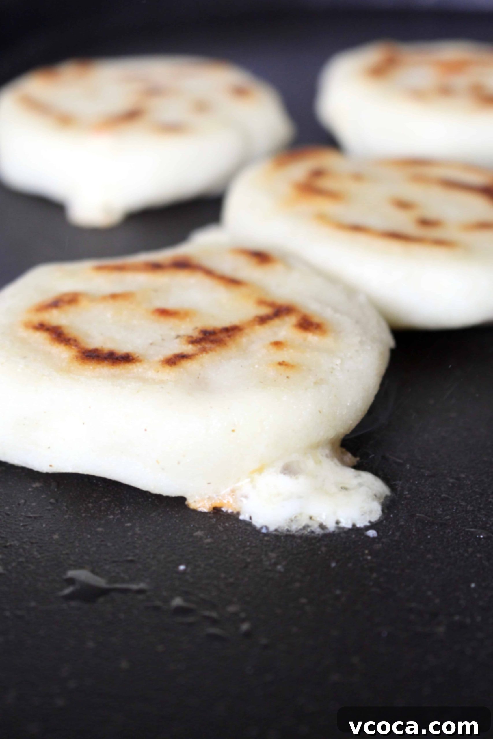 Melted cheese oozing from inside an arepa as it cooks on a black griddle, showcasing a delicious cheese-stuffed arepa.
