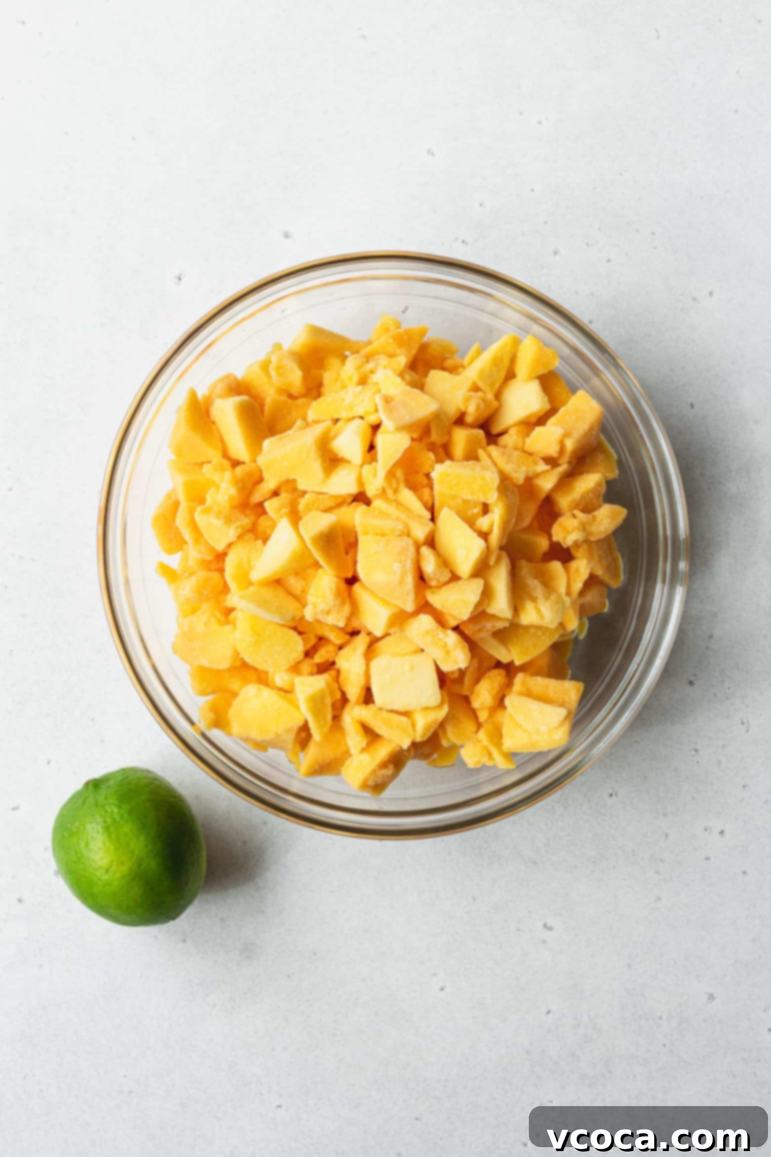 Easy Two Ingredient Mango Sorbet 3 A bowl of freshly cut frozen mango chunks, perfectly prepped for blending into sorbet, accompanied by a whole ripe lime on a light surface