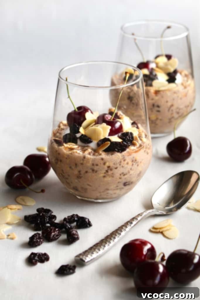 Two glass tumblers filled with creamy cherry almond overnight oats, garnished with fresh cherries, dried tart cherries, and toasted sliced almonds scattered around for a vibrant, appealing breakfast display.