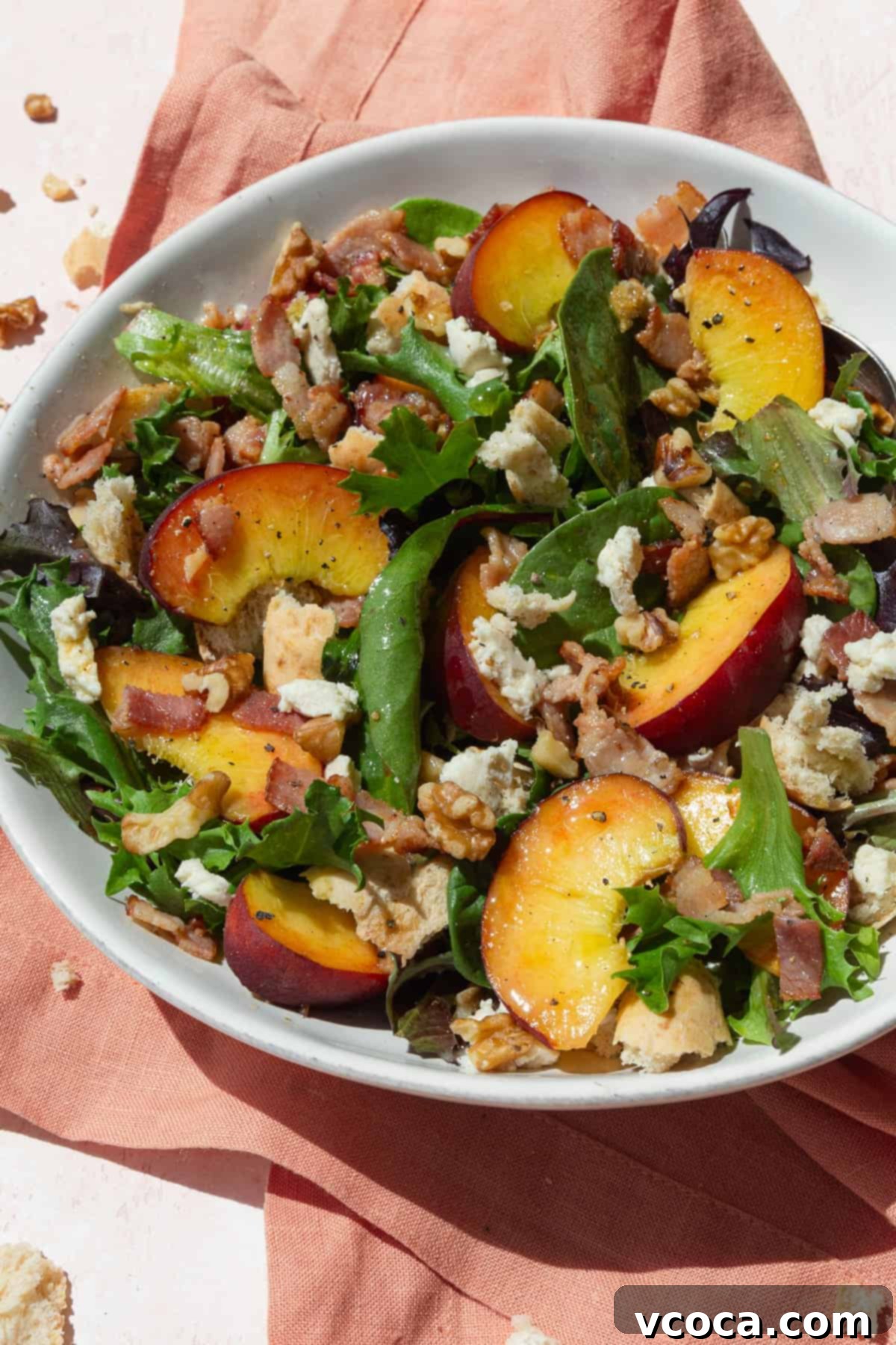 Close-up of a vibrant peach panzanella salad with baby greens, creamy cheese, and toasted walnuts in a white bowl on a pink napkin.