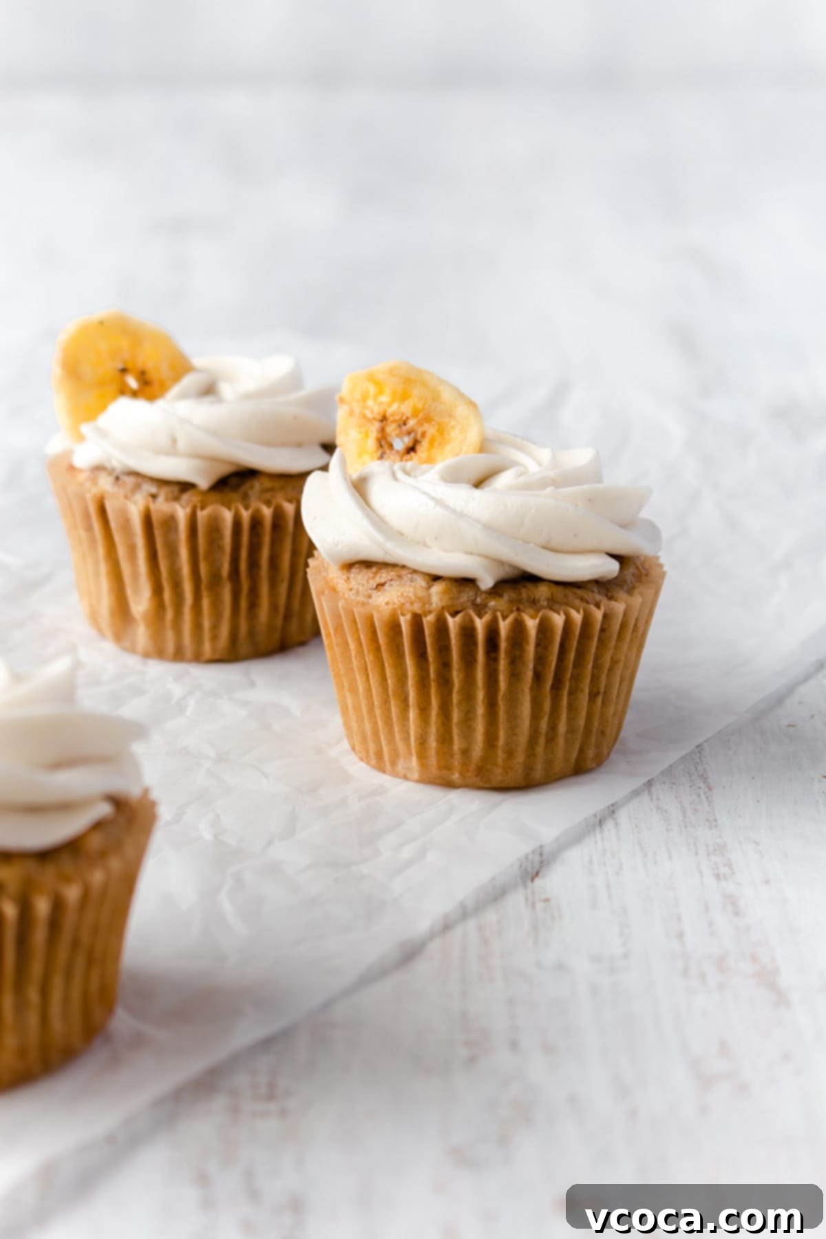 Two banana cupcakes on a piece of parchment paper with another cupcake off to the side, showcasing their fluffy texture and simple elegance.